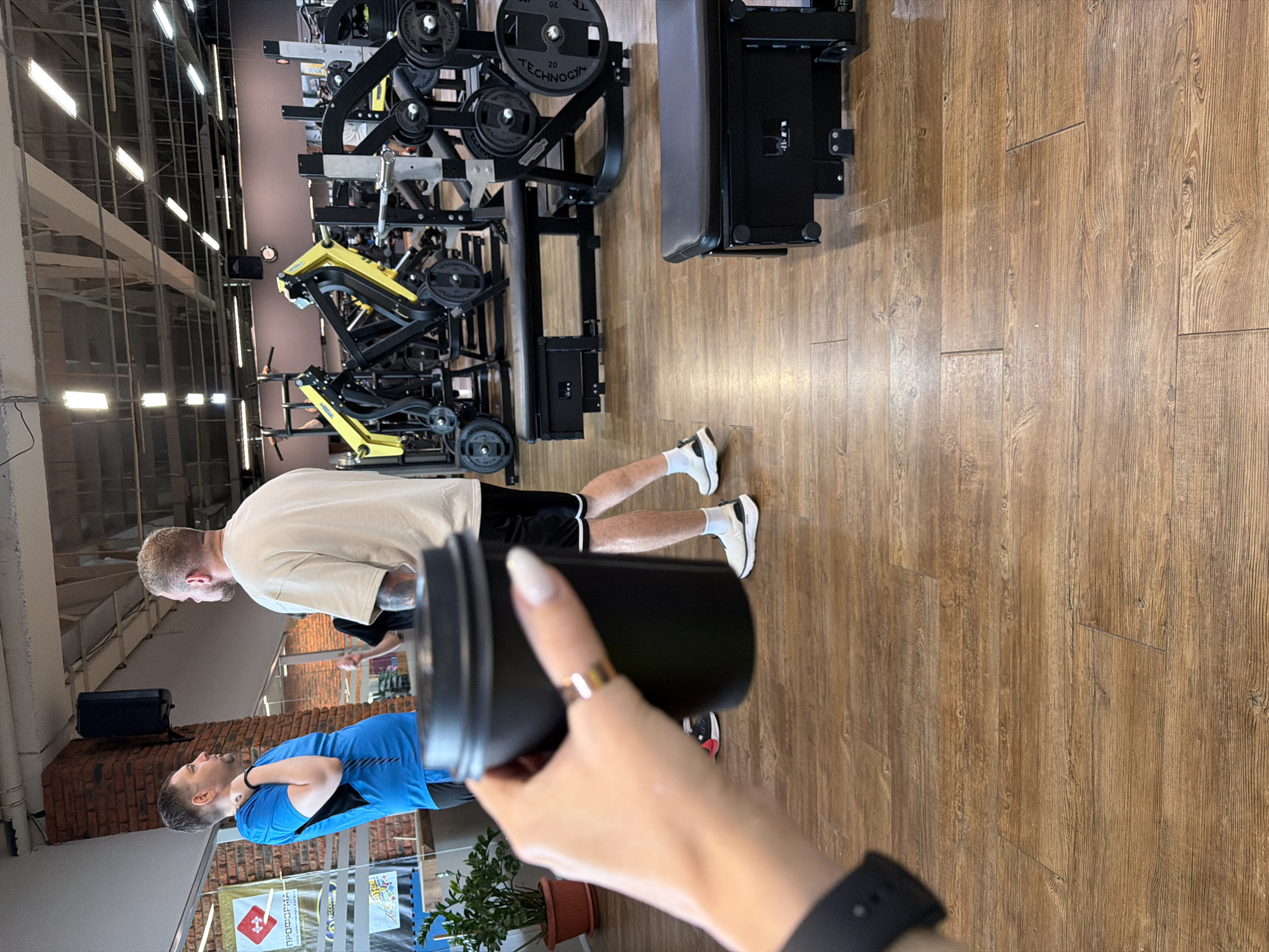 The image depicts the interior of a fitness center, possibly in Chaikovsky, Russia. Two men are visible: one wearing a blue shirt and the other in a white shirt and black shorts. The man in the white shirt is further away from the camera and wearing white sneakers with white socks. The man in the blue shirt is positioned closer to the camera and is wearing a black wristband on his left arm. A hand holding a black shaker is prominently featured in the foreground, obscuring some of the view. A black watch is visible on the wrist of the person holding the shaker. Various pieces of exercise equipment, including black weight machines, are positioned in the background. The floor is made of wood. A potted plant is present near the lower left corner.