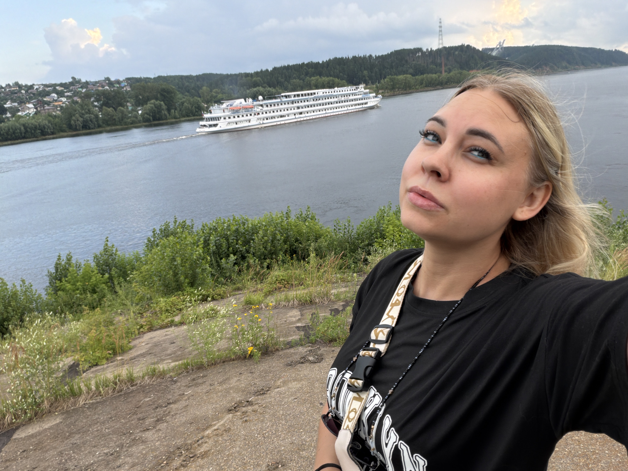 A young woman with blonde hair and blue eyeshadow poses for a selfie on a dirt embankment overlooking a wide, calm river. She is wearing a black t-shirt with a graphic design and a camera strap with a beaded necklace. In the background, a large white riverboat is cruising down the river. On the opposite bank, a small town with red-roofed houses is nestled amongst green trees. Further in the distance, a tall industrial tower stands against a cloudy sky. The scene suggests a scenic view of Chaikovsky, Russia, likely near the Kama River.