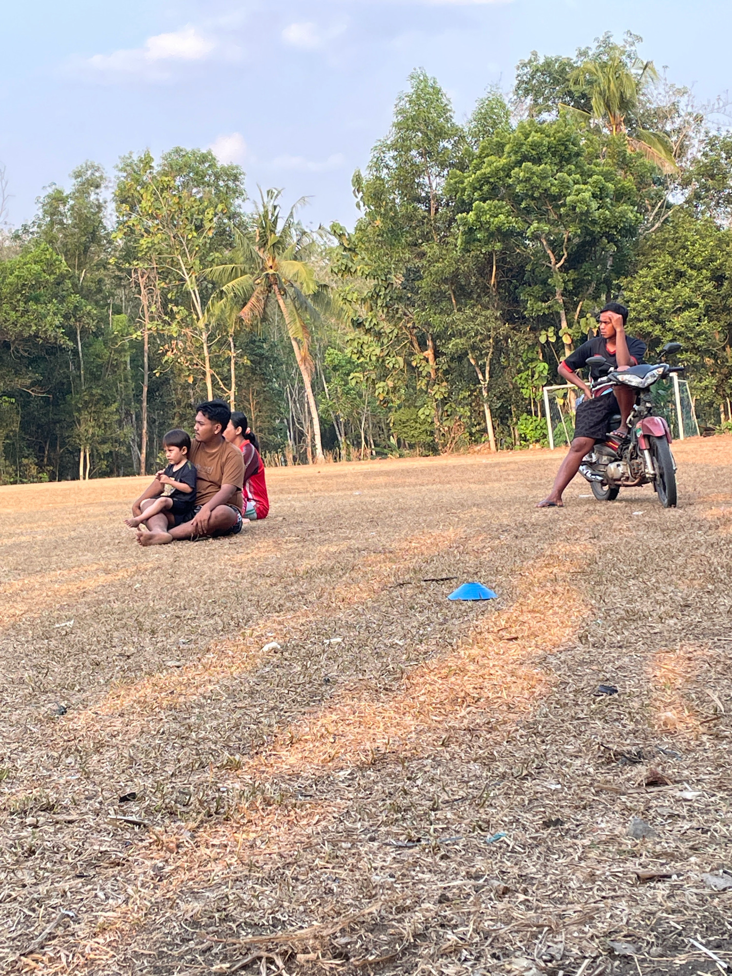 In Kagungan Ratu, Indonesia, on a dry, grassy field, a young boy sits on the lap of a man. The man is wearing a brown t-shirt and dark shorts, and the boy is in a black t-shirt and shorts. They are seated on the ground, with another person in a red shirt behind them. Further in the background, a young man is seated on a red motorcycle, resting his head on his hand. Trees and a soccer goal frame the scene, under a lightly clouded sky.