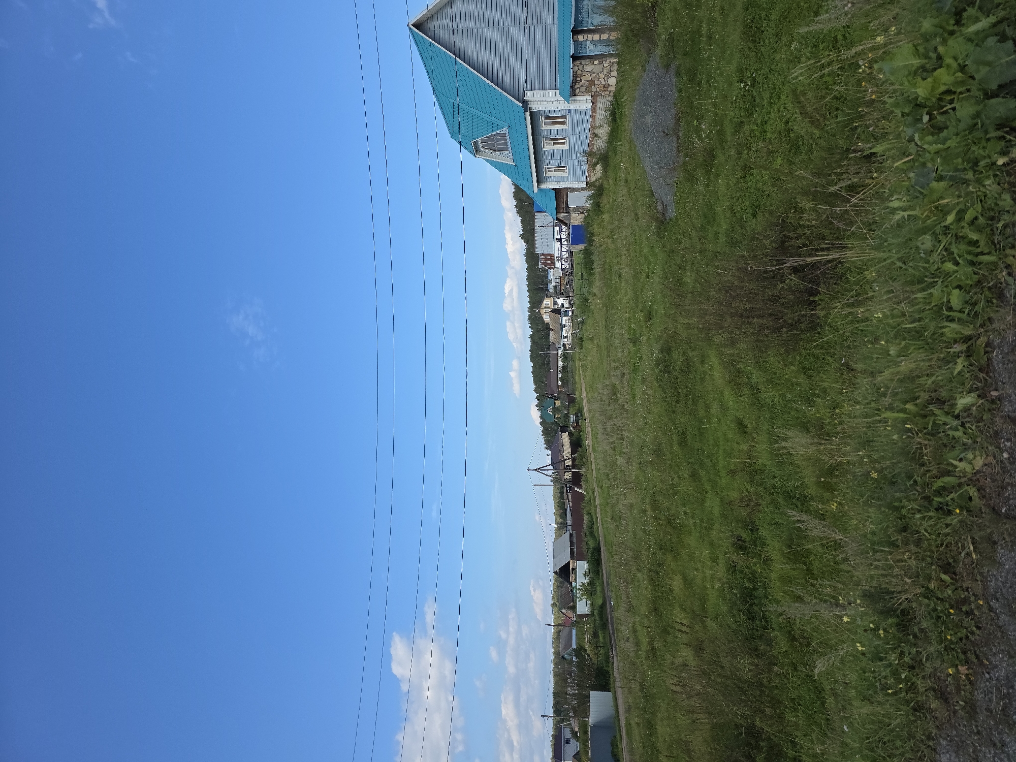 The image depicts a residential area in Khalitova, Russia, under a clear blue sky with scattered clouds. A prominent blue-roofed house with light blue siding and a dormer window is situated on the right side of the frame, bordered by a stone and brick foundation. To the left of this house, a dirt road runs through a grassy field dotted with various structures, including fences, smaller houses, and what appear to be sheds or outbuildings. Power lines are visible strung across the upper portion of the image, crossing over the landscape and the houses. The foreground consists of a grassy area with wildflowers and weeds, with a gravel surface at the very bottom. The overall scene suggests a rural or semi-rural setting.