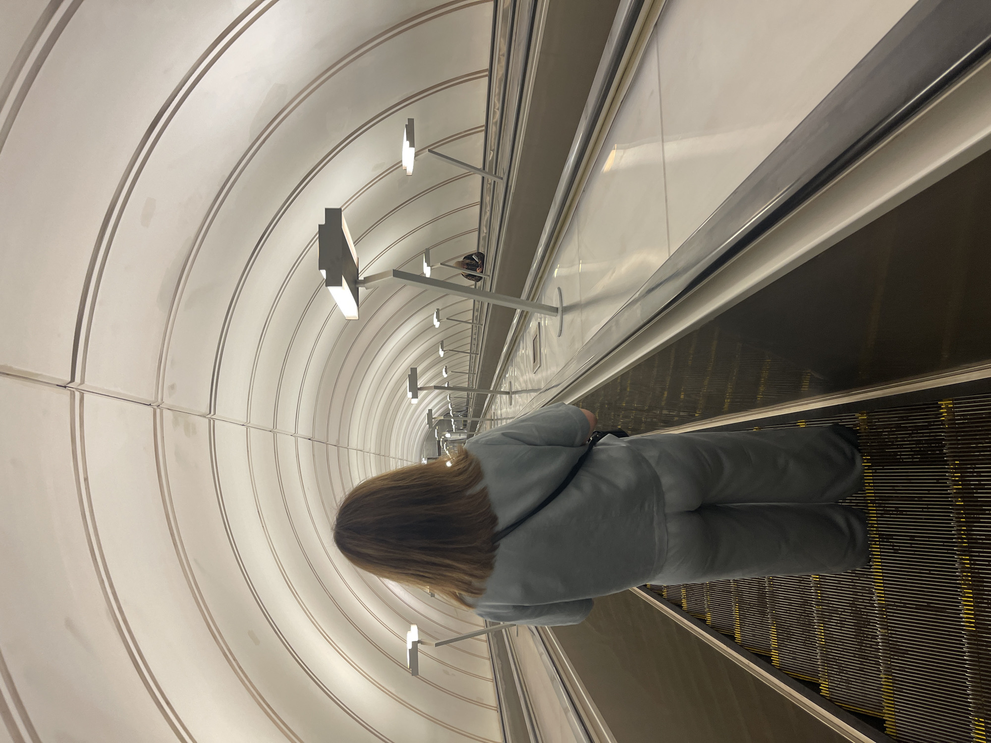 A person stands on an escalator within the Moscow Metro. The escalator ascends through a tunnel with a white, vaulted ceiling, illuminated by a row of modern light fixtures. The person is wearing a light-colored tracksuit and faces away from the camera. The escalator itself has metallic sides and treads.