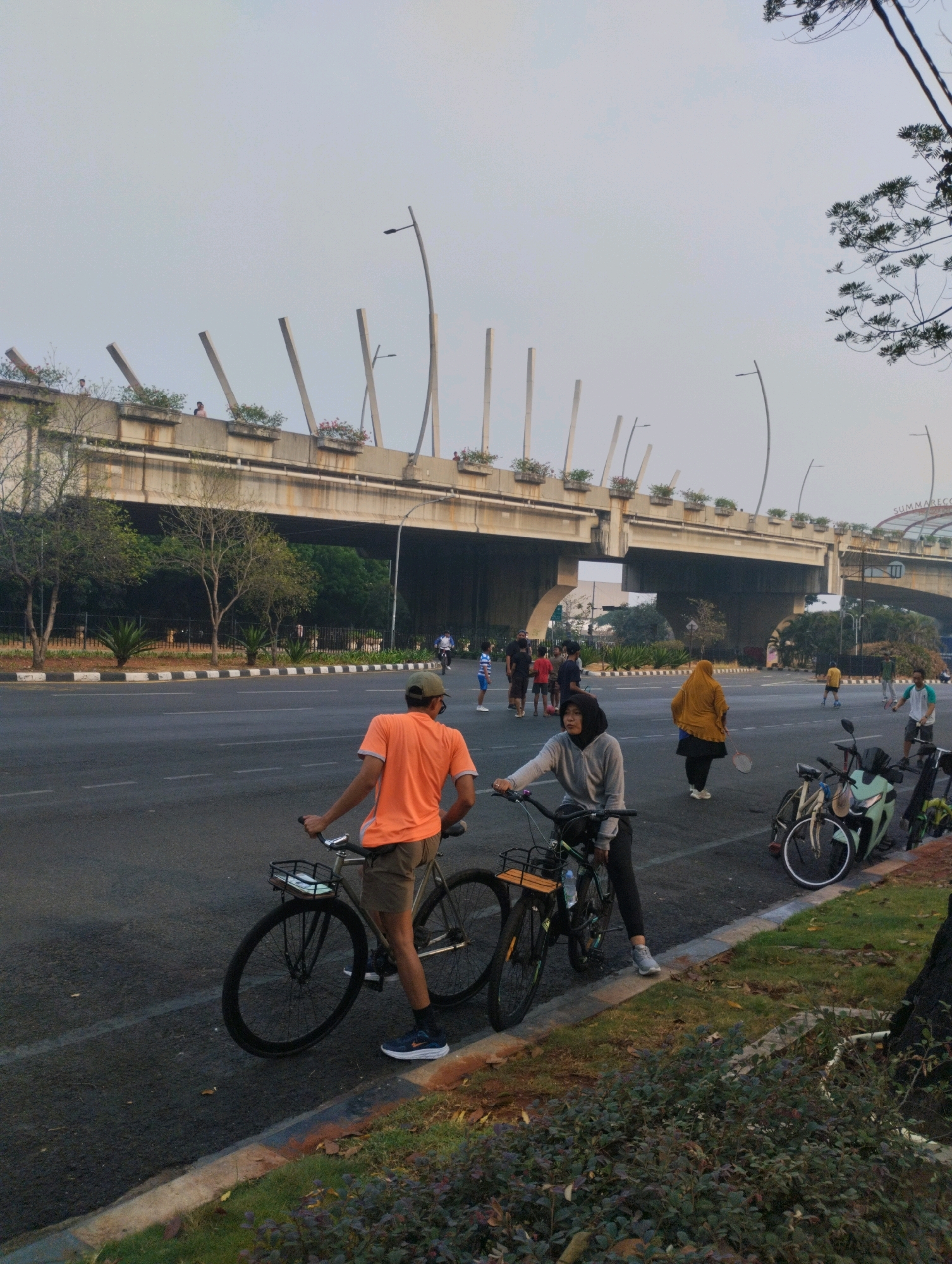 In Bekasi, Indonesia, an outdoor scene unfolds on a wide asphalt road, likely a thoroughfare for recreational activities. The sky is overcast, casting a diffused light over the environment. In the foreground, two individuals are prominently featured. A person in an orange t-shirt and khaki shorts stands with their back to the camera, holding onto the handlebars of a bicycle. To their right, a person wearing a grey hooded jacket and black leggings is also by a bicycle. Further into the background, a group of people are engaged in various activities. Some are playing badminton, as evidenced by rackets and shuttlecocks. Others are walking or cycling, with one individual in a mustard-colored hijab walking with a badminton racket. Several bicycles and a scooter are parked along the side of the road. In the background, a large overpass, characteristic of urban infrastructure, spans across the scene, with decorative planters and lamp posts lining its top. Trees and foliage line the median and the edge of the road, adding a touch of nature to the urban landscape. The overall impression is one of a lively community gathering, with people enjoying outdoor activities in a city setting.