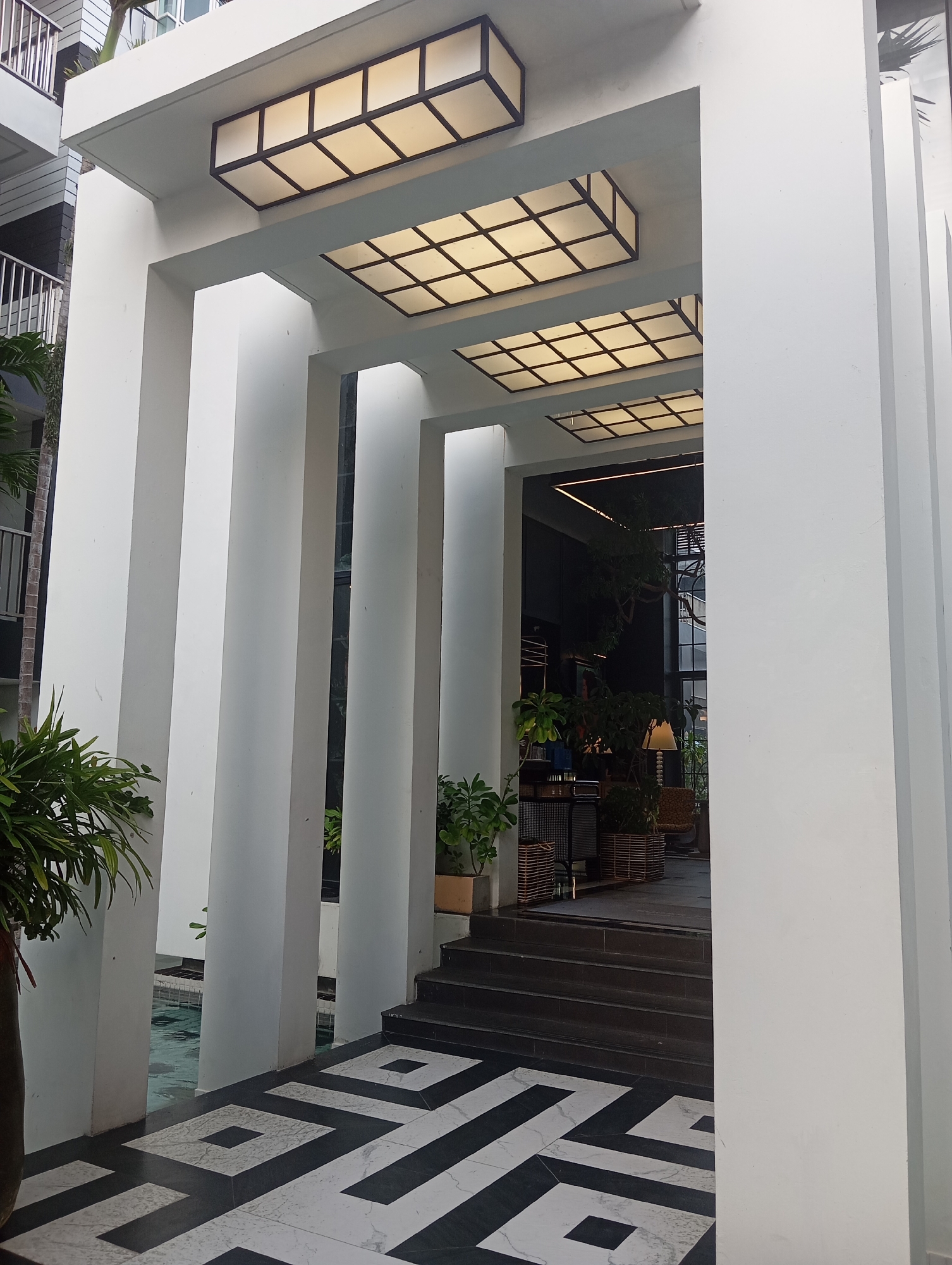The image shows an outdoor entrance to a building with a modern architectural design. The entrance is framed by large white pillars and a white overhead structure with recessed rectangular lights. The floor leading to the entrance features a black and white geometric pattern. To the left of the entrance, there is a glimpse of a swimming pool. In the background, visible through the entrance, is an indoor space with plants, furniture, and a staircase. The scene is well-lit, suggesting it is daytime. The location is identified as Karon, Thailand.