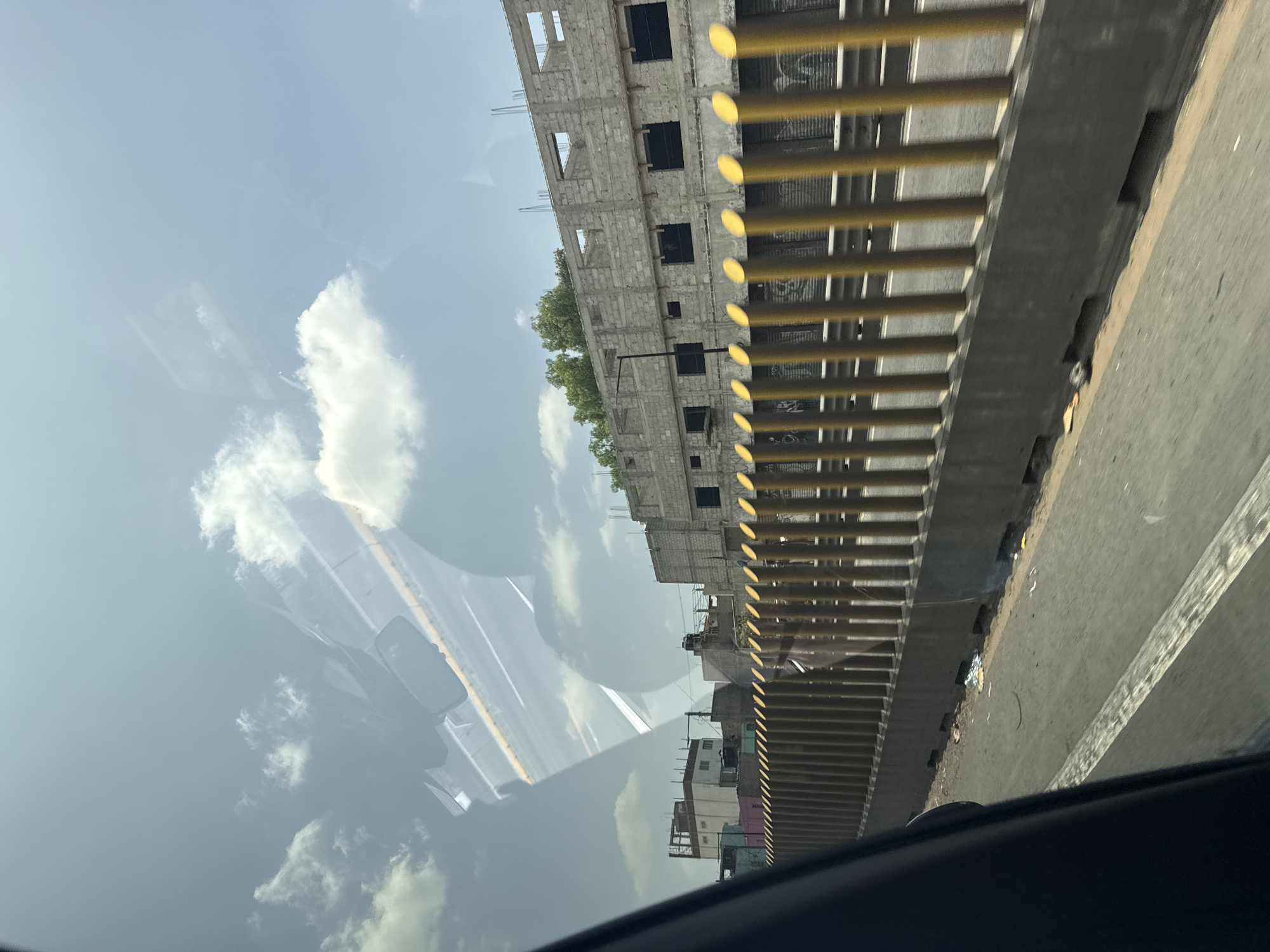 The image depicts a view from inside a vehicle, possibly a car, in Valle de Chalco Solidaridad, Mexico. Through the window, the sky is visible, with light clouds and a reflection of the vehicle's interior. Outside, alongside the road, there is a concrete barrier painted with horizontal yellow and grey stripes. Beyond the barrier, there's an unfinished, multi-story building made of concrete blocks with visible window openings. Further in the distance, there are other buildings, presumably residential, and some sparse vegetation. Litter and debris are visible on the side of the road. The image is taken at an oblique angle.