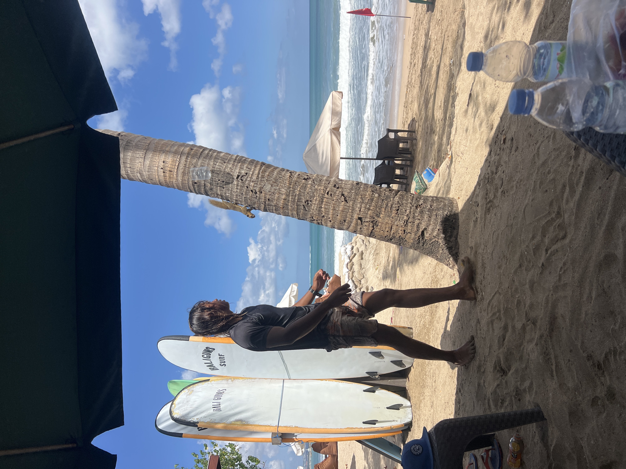 A man stands on a sandy beach in Kuta, Indonesia, looking out at the ocean. He is wearing a dark t-shirt and patterned shorts. Behind him, three surfboards stand upright, two with "BALIGUNS SURF" printed on them. A large palm tree trunk is positioned in the foreground, casting a shadow on the sand. Beyond the man and surfboards, beach chairs and an umbrella are visible near the water's edge, where waves are breaking on the shore. The sky is blue with scattered white clouds.