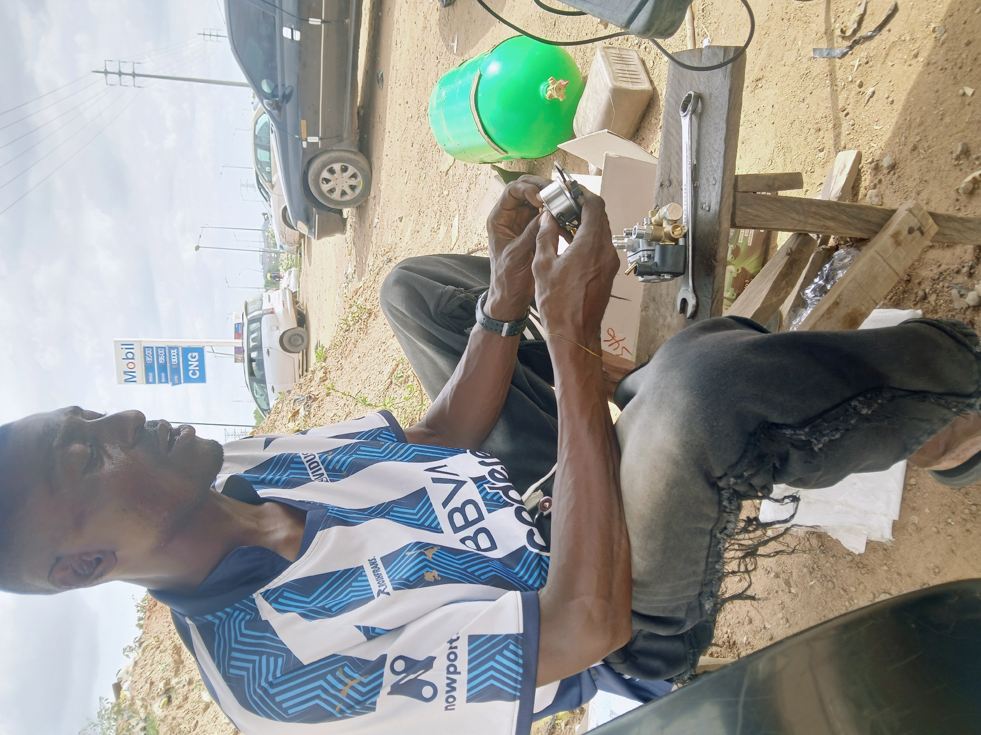 An African man is sitting outdoors, seemingly working on or assembling a small engine part or device. He is wearing a blue and white soccer jersey and dark-colored pants. He is sitting on a makeshift wooden stool or small table. A wrench and a green gas cylinder are on the table beside him, and an open box suggesting it contains the part he is working on. In the background, a Mobil gas station sign is visible, along with cars parked along the side of the road. Based on the style of the Mobil sign and general environmental details, the location is likely in Abuja, Nigeria. The man is holding a metallic object in his hands and appears focused on it.