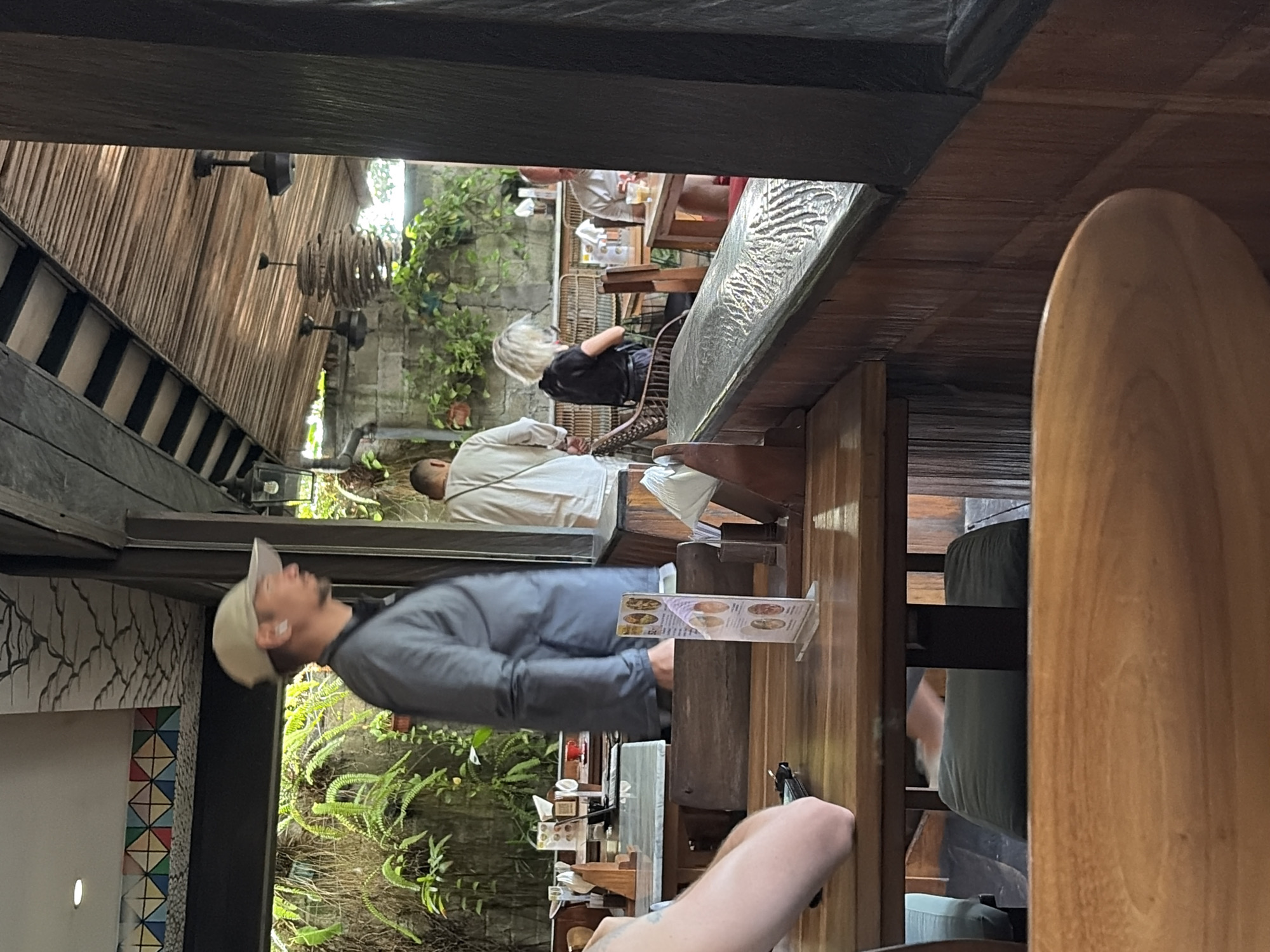 The image depicts the interior of a restaurant in North Kuta, Indonesia. In the foreground, a wooden table is visible, with a person's arm resting on it. Behind this table, another wooden table is set with a menu stand. A man wearing a grey long-sleeved shirt and a light-colored baseball cap is standing and looking to the right. Further into the background, several other patrons are seated at tables. One person in a white garment is visible from the back, and a woman with blonde hair is seated. A man with a light-colored shirt is also seated at a table with a drink. The environment is open-air, with lush greenery visible through an opening and a rustic wooden ceiling with fans. The overall atmosphere suggests a casual dining setting.