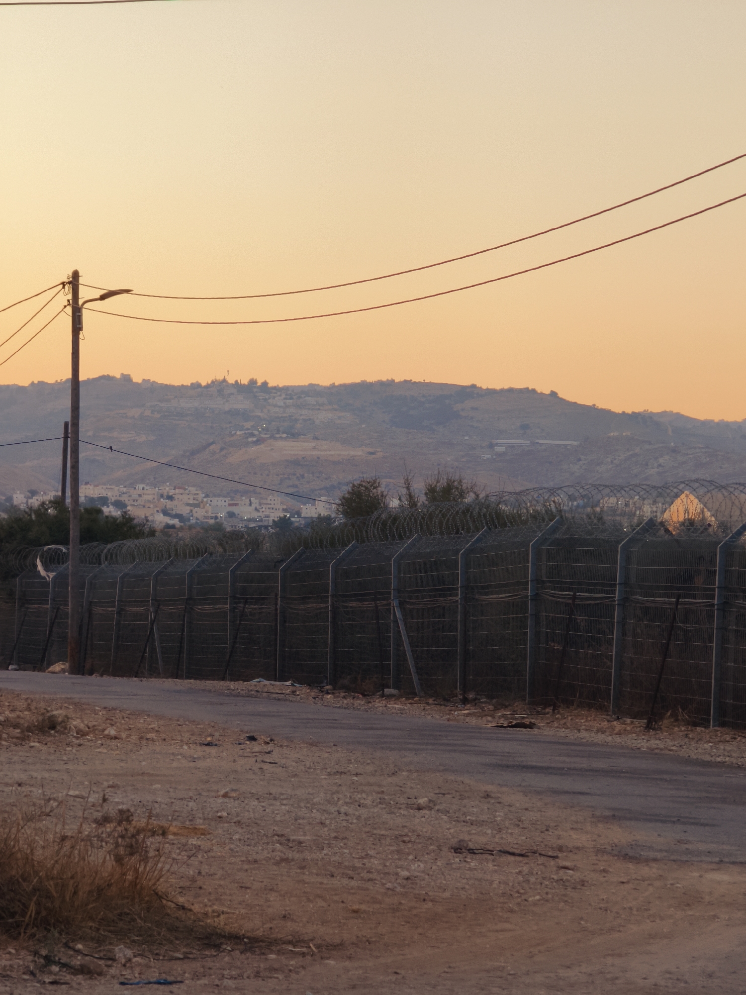 The image captures a rural road in the Lachish Regional Council, Israel, at sunset. A chain-link fence topped with barbed wire runs along the right side of the road, separating it from a treeline and a distant settlement on a hillside. The sky is a soft orange, indicative of dusk, with power lines stretching across the frame. The foreground consists of a dirt and gravel road, with dry grasses and sparse vegetation on the left. In the background, a town or village with light-colored buildings is visible on the undulating landscape. The overall scene suggests a border or controlled area, given the substantial fencing.