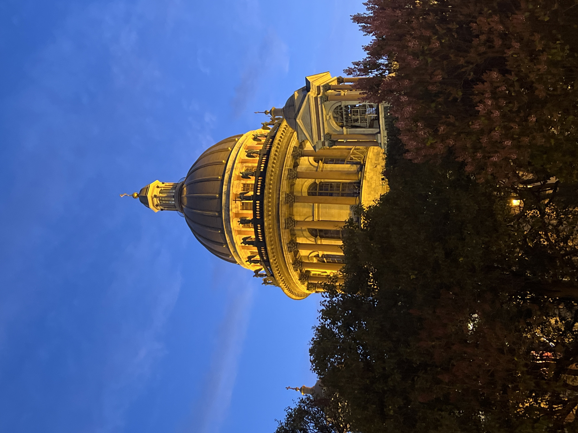The image shows a low-angle view of St. Isaac's Cathedral in Saint Petersburg, Russia, at dusk or night. The cathedral's dome, illuminated in warm tones, is the main focus. Part of the architectural structure below the dome, featuring columns and windows, is visible. To the right of the cathedral, trees with dark foliage partially obscure the view. The sky is a deep blue with some light cloud cover. No people are visible in the scene.