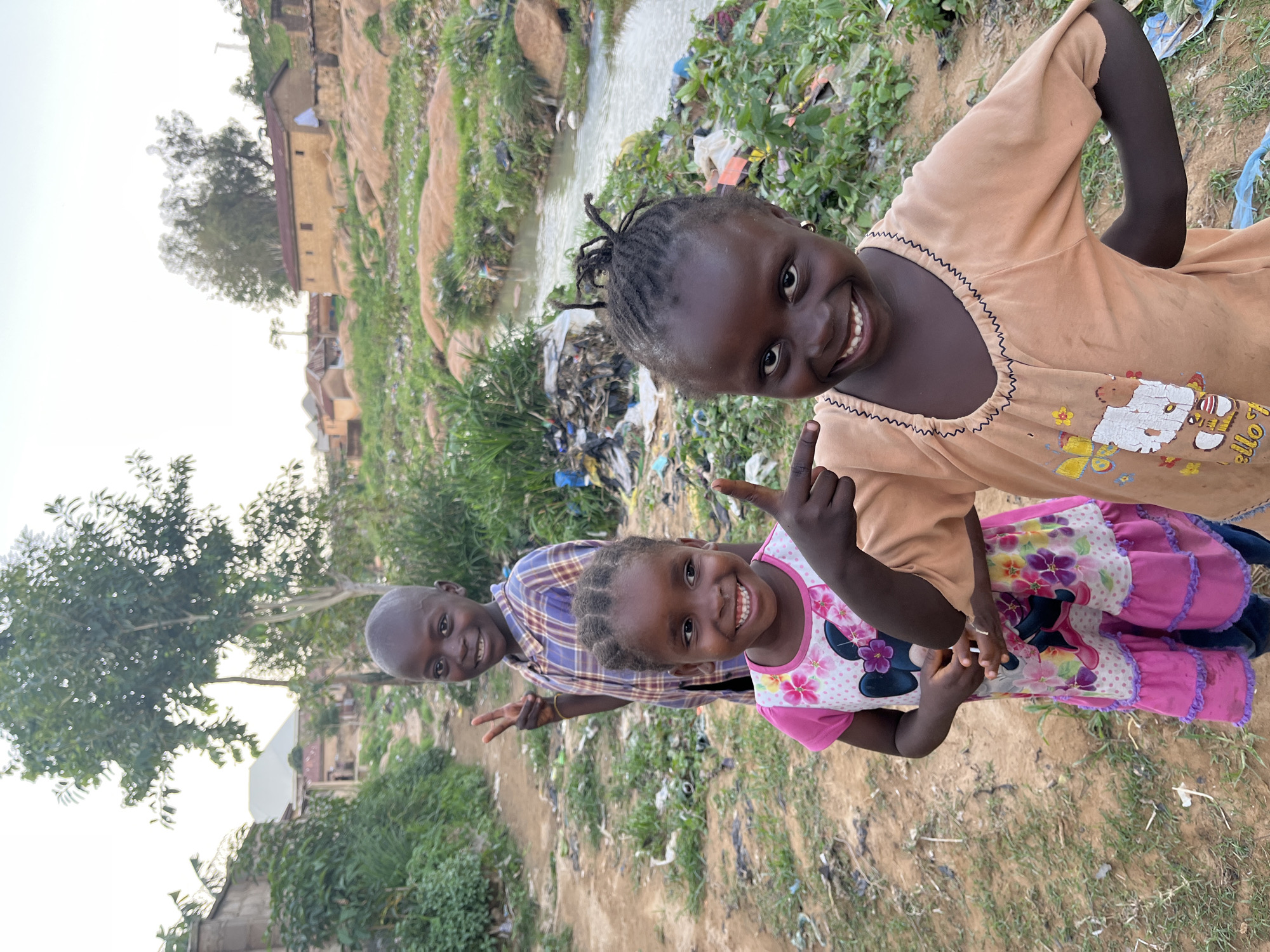 The image shows three children smiling and posing for a photo. Two girls are in the foreground, and a boy is behind them. The girl on the right is wearing a peach-colored shirt with a graphic of Mickey Mouse. The girl in the middle is wearing a pink dress with a floral pattern. The boy on the left is wearing a plaid shirt. They appear to be in an outdoor setting with a stream and some vegetation in the background. There is also evidence of trash and debris in the background, suggesting a less developed area. The lighting suggests it is daytime. The children's smiles and gestures indicate a happy and playful mood.