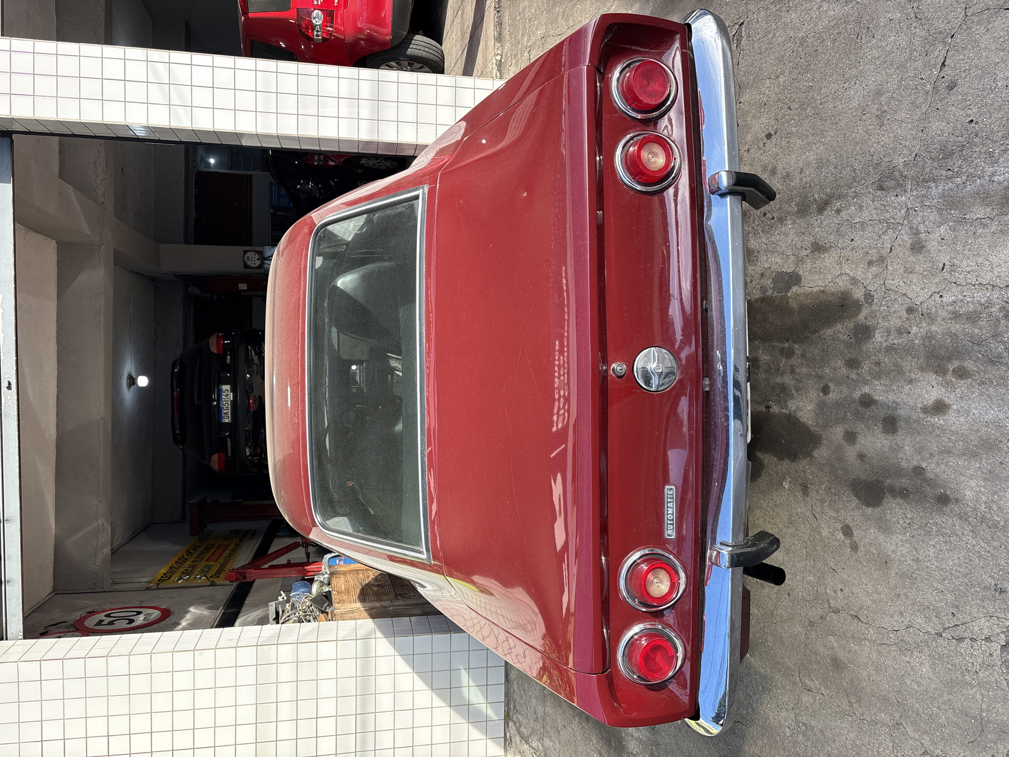 The image shows the rear end of a red vintage car parked inside a garage in São Paulo, Brazil. The car, a Chevrolet Opala coupe, is the primary subject, with its distinctive taillights and chrome bumper prominent. Behind it, other vehicles, including a black Peugeot with a license plate ending in "G5H45" and a red SUV, are visible in the garage. The garage appears to be a workshop, with tools and equipment, including a car lift, present. The walls are tiled with white squares, and a speed limit sign showing "50 km/h" is mounted on the left wall. The ground is a cracked concrete surface with oil stains. The lighting suggests it is daytime.