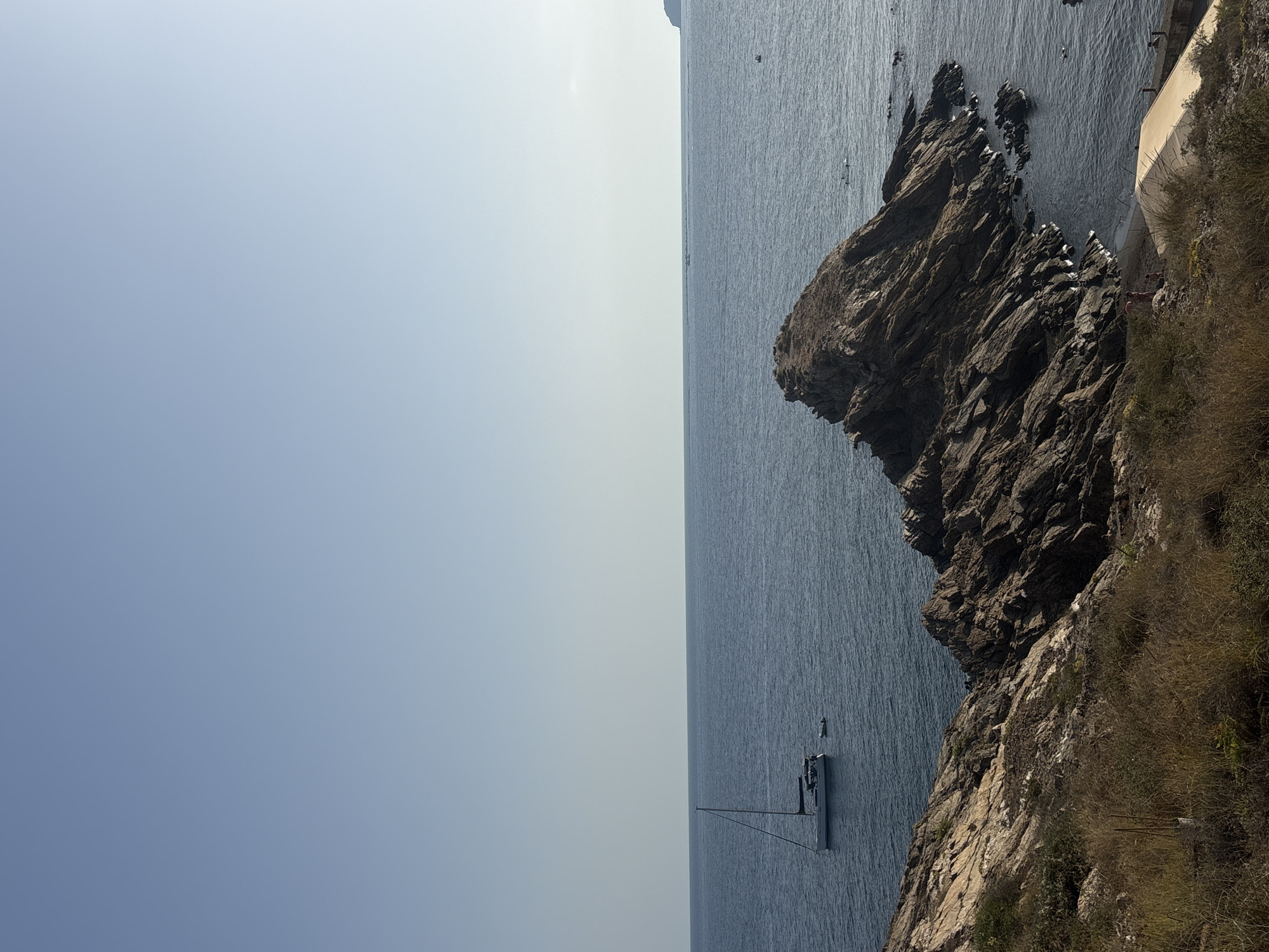 The image depicts a coastal scene in La Unión, Spain, characterized by a clear sky and calm sea. In the foreground, a rocky outcrop covered in dry grass and brush slopes down towards the water. A large, jagged rock formation juts out into the sea, its dark, striated surface contrasting with the blue water.
Further out in the sea, a white sailboat with a single mast is visible, appearing to be at anchor. A small inflatable dinghy is tied to the stern of the sailboat. A few other distant boats can be seen on the horizon.
On a small sandy beach nestled between rocks, a few people are visible. One person appears to be standing on the beach, and another is further down, near a light-colored sunshade or awning. Several swimmers are dotted in the water near the rocky shore. The overall atmosphere is one of peaceful coastal recreation.