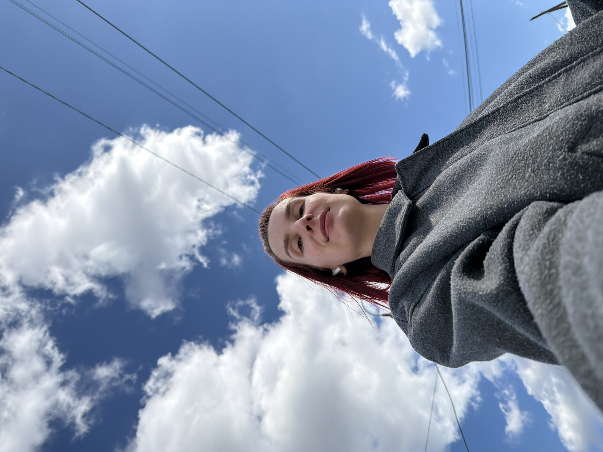 A low-angle outdoor shot captures a young person with reddish hair, dressed in a gray, textured jacket, looking directly towards the camera. The individual, visible from the chest up, has a faint smile and appears to be wearing white wireless earbuds. Their gaze is slightly upward, aligning with the camera's perspective.
The background is dominated by a clear, vibrant blue sky dotted with scattered white cumulus clouds. Several thin, dark power lines (or similar utility wires) stretch horizontally and diagonally across the upper portion of the sky, adding an urban or suburban element to the otherwise natural backdrop. The bright lighting suggests it is daytime, under fair weather conditions. The composition creates an expansive feel, with the person positioned against a vast sky. No visible text is present in the image. This scene takes place in Chaikovsky, Russia.