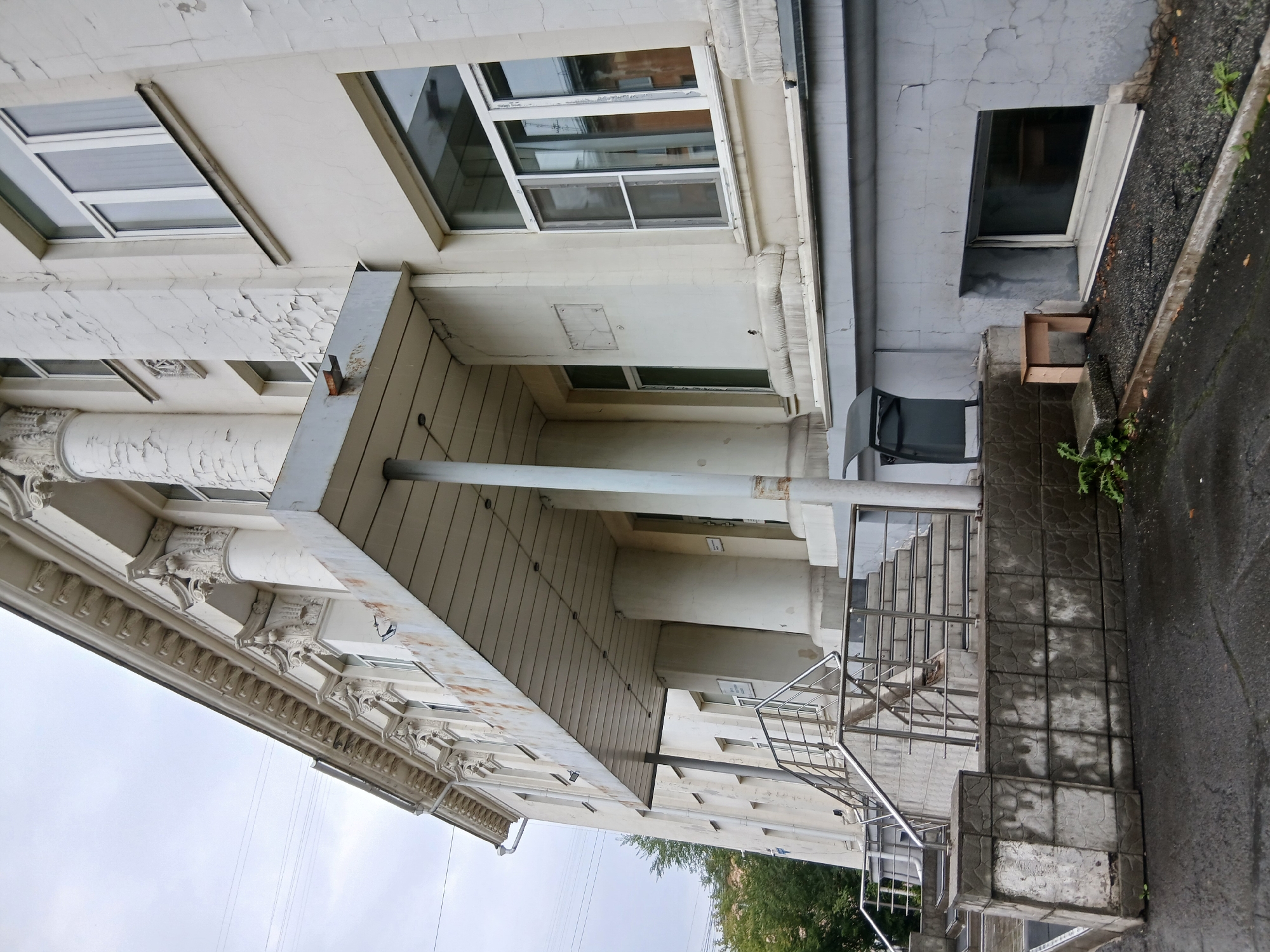 The image shows the exterior of a weathered, pale yellow, multi-story building, likely of institutional or governmental use, with architectural elements such as classical columns and ornate cornices. The paint is peeling in many areas, revealing cracks and discoloration. A covered entrance with a metal awning is situated above a set of concrete stairs with a metal handrail. The stairs lead up from a wet, asphalt ground. To the left of the stairs is a low, concrete retaining wall covered in textured tiles, some of which are stained. To the right of the stairs, near the foundation, is a small, light-colored wooden stool and a dark gray trash receptacle. A basement-level window is visible on the right side of the image, set into a cracked, gray-painted foundation wall. The sky is overcast, and trees are visible on the left side of the building. The overall impression is one of age and disrepair.