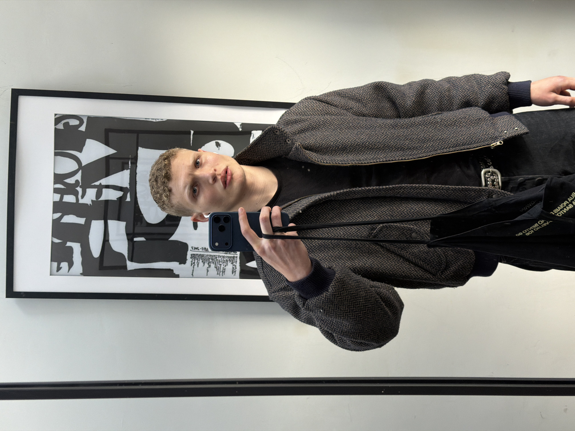 The image shows a fair-skinned person with short, curly blonde hair taking a selfie in a mirror. They are wearing a black t-shirt and a brown and grey herringbone patterned jacket with navy blue wrist cuffs. The person is holding a blue smartphone. A black tote bag is slung across their body. A framed black and white abstract print hangs on the wall behind the person. The location appears to be indoors with white walls. A black bar runs along the bottom of the frame, suggesting it's part of a mirror or fixture. There is no indication of the city or country of location within the frame.