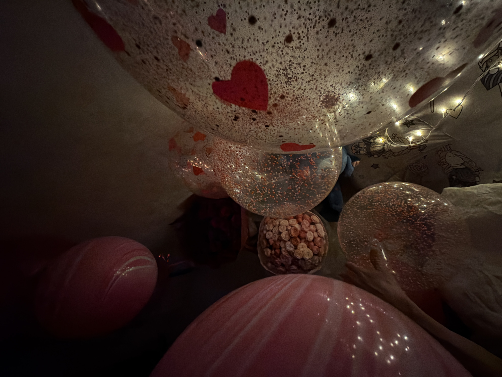 The image depicts an interior scene, possibly a room decorated for a special occasion. Dominating the view are several balloons of varying sizes and designs. Some balloons are transparent with red heart patterns, while others are pink with white swirls. A cluster of small, round objects, possibly small flower arrangements or candy, sits in a clear container. In the background, string lights are visible, contributing to the festive atmosphere. The room appears dimly lit, with a casual and intimate ambiance. The location, Chaikovsky, Russia, is provided.