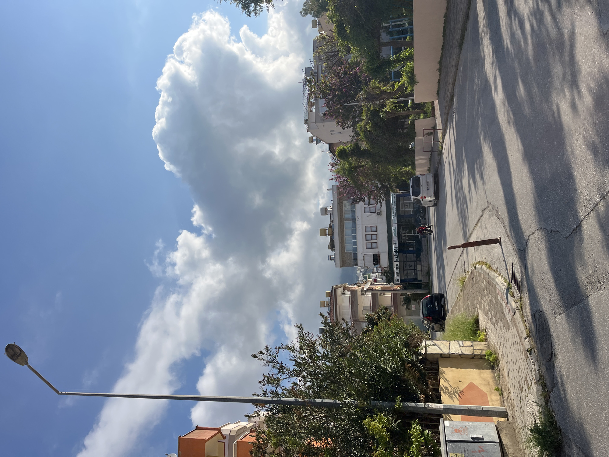 The image depicts a street scene in Alanya, Turkey. The street is paved and flanked by buildings of varying heights and architectural styles. Lush vegetation, including trees and shrubs, lines the street and obscures some of the buildings. A street lamp stands prominently in the foreground. In the background, there are large cumulus clouds against a blue sky. A car is parked on the side of the road. A motorcycle is moving on the street.