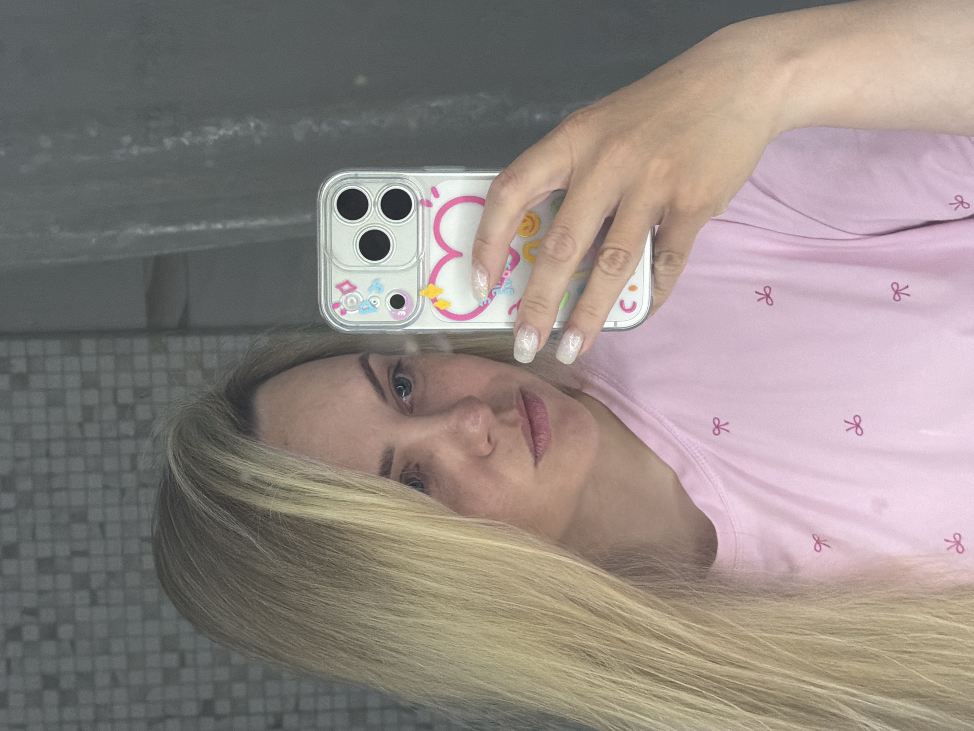 A fair-skinned, light-haired woman takes a selfie in what appears to be a bathroom. She holds a cell phone with a decorative case, the back of the phone visible with its three camera lenses. She wears a light pink top with small, dark pink bow patterns. The background consists of a tile wall and a reflection in a mirror. The location in Votkinsk, Russia is unknown.