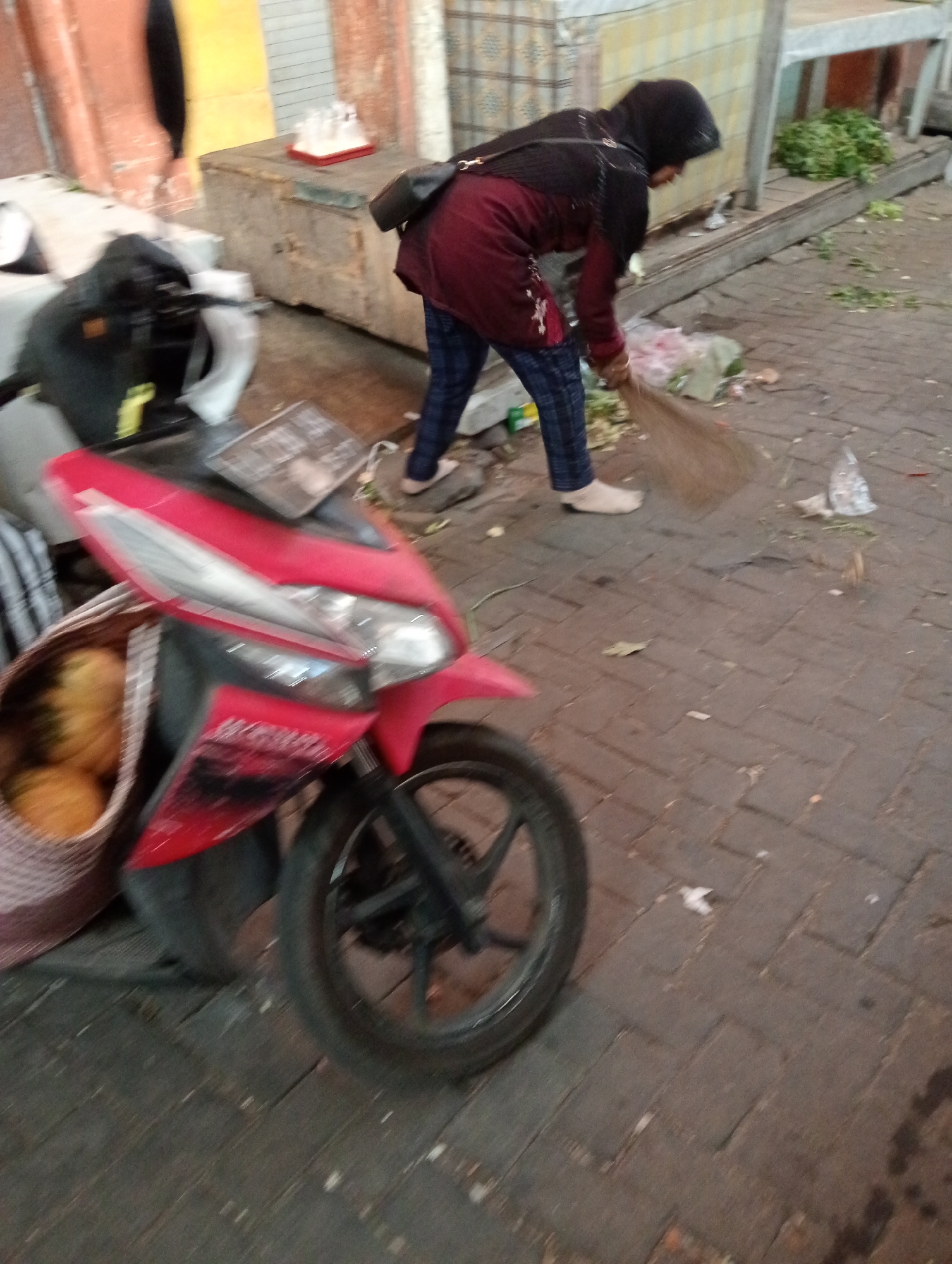 In Grabag, Indonesia, a person wearing a black hijab and a maroon jacket with blue and white checkered pants is sweeping the ground with a broom. The person is standing on a brick-paved surface, and there is debris scattered on the ground, including some green leaves and plastic bags. To the left of the person, a red and black motorcycle is parked, with a basket containing yellow fruit attached to its front. In the background, there is a counter with some items on it, and a wall with orange and yellow paint. The scene appears to be outdoors, possibly in a market or street setting.
