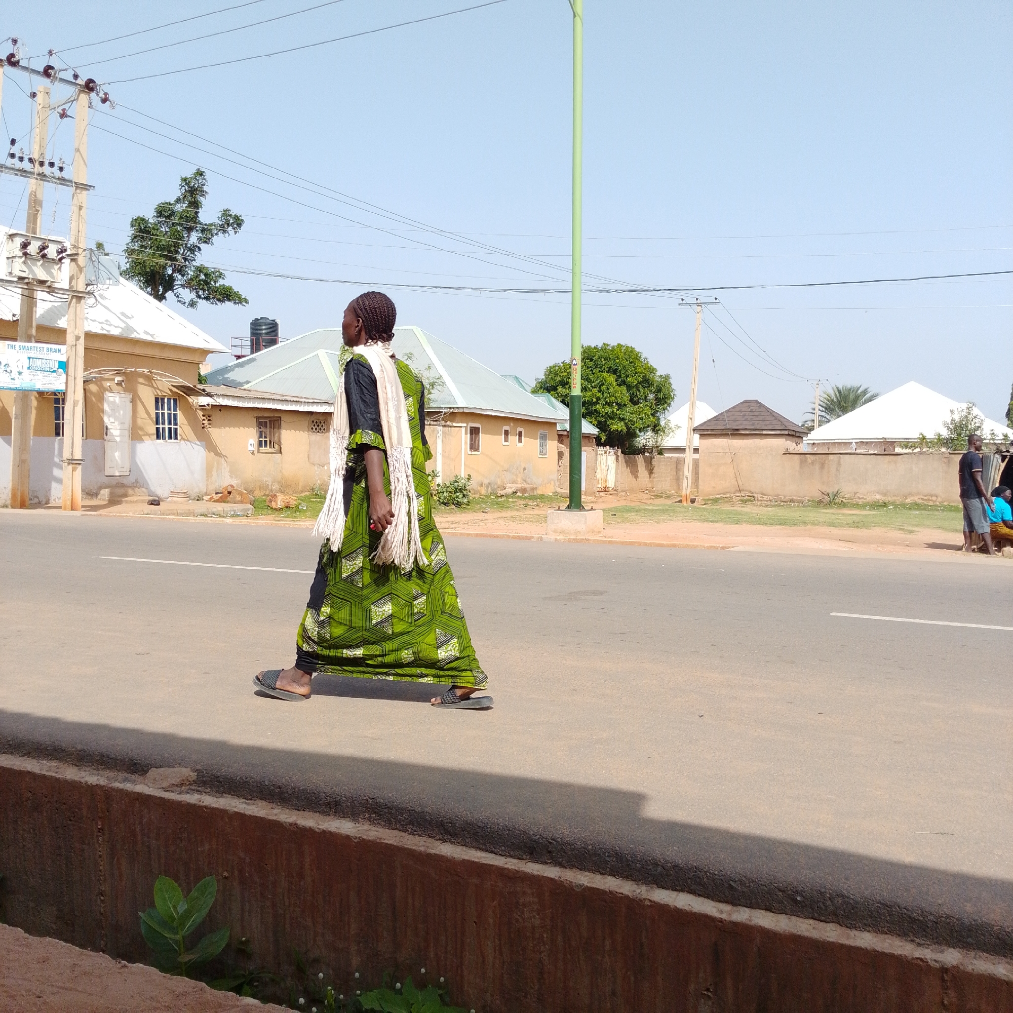 A woman walks along a paved road in Gombe, Nigeria. She is wearing a long, green, patterned dress with black leggings underneath and a light-colored scarf draped over her shoulders. She is also wearing black sandals. The woman is walking from left to right in the frame, with her body angled slightly away from the camera.
The scene is an outdoor, daytime setting under a clear, pale blue sky. Buildings with light-colored walls and pale roofs are visible in the background, suggesting a residential or commercial area. Trees and utility poles with numerous wires are also present. A tall, light green pole with some signage is situated to the right of the woman. The road has a white line marking the center. In the distance, two men and a woman are standing near a structure, and another person is walking away from the camera.
The overall atmosphere appears to be calm and ordinary, with no indication of any particular event or strong emotion. The lighting suggests it is daytime, likely late morning or early afternoon due to the bright sunlight and distinct shadows. The presence of a water tower on one of the buildings provides a clue to the location's infrastructure. A sign is visible on the left side of the frame, but the text is too small to be legible.