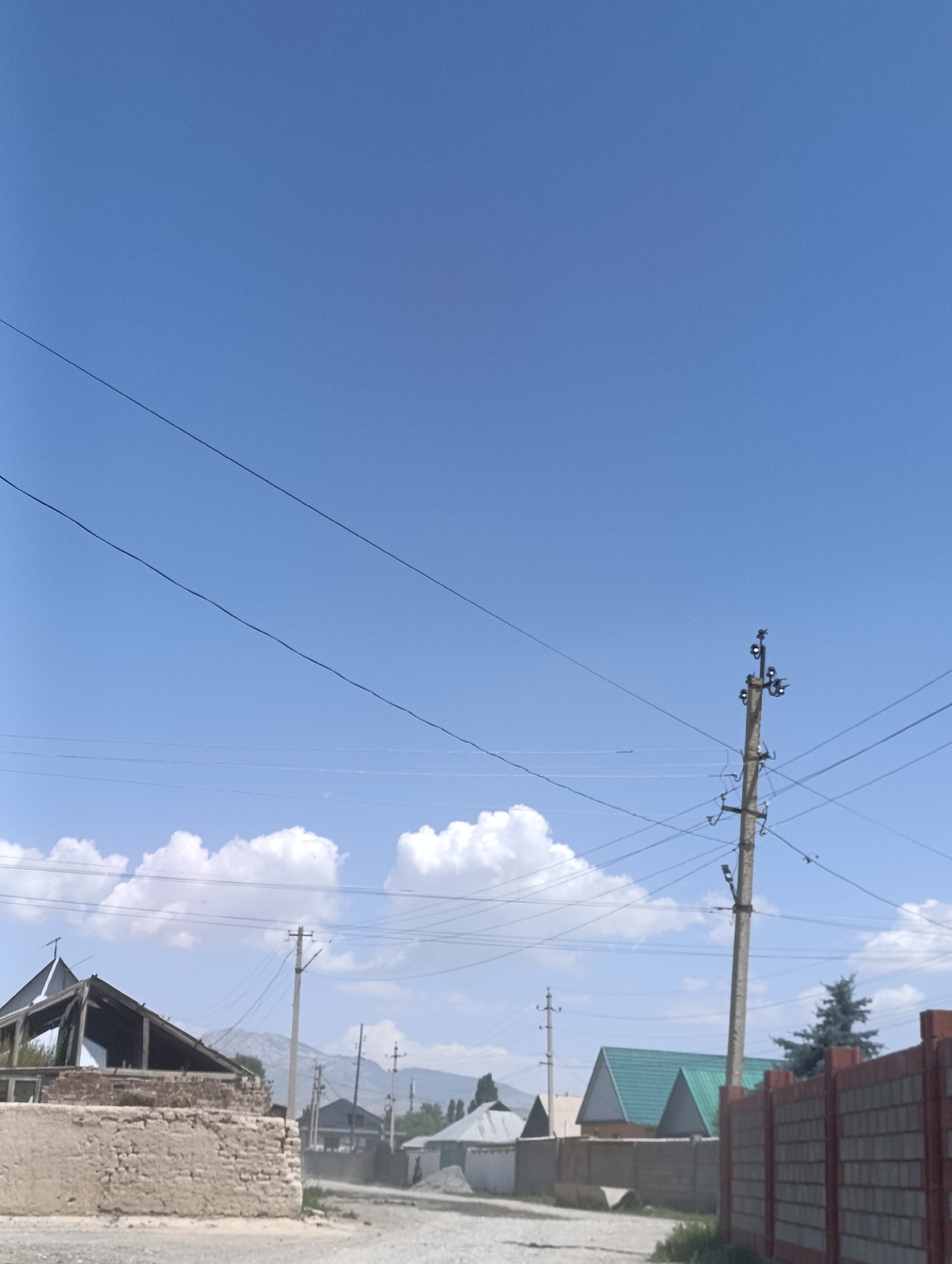 The image depicts a street scene in At-Bashy, Kyrgyzstan, under a clear blue sky with scattered clouds. In the foreground, to the left, is a partially constructed or damaged building with a brick base and exposed wooden framing and roof structure. Further down the street, utility poles with wires crisscross the sky. Several houses with various roof designs are visible, including one with a distinctive green, pitched roof. A fence made of concrete blocks runs along the right side of the image. The ground appears to be a dirt or gravel road. In the background, mountains are visible on the horizon. No people or animals are present in the frame.