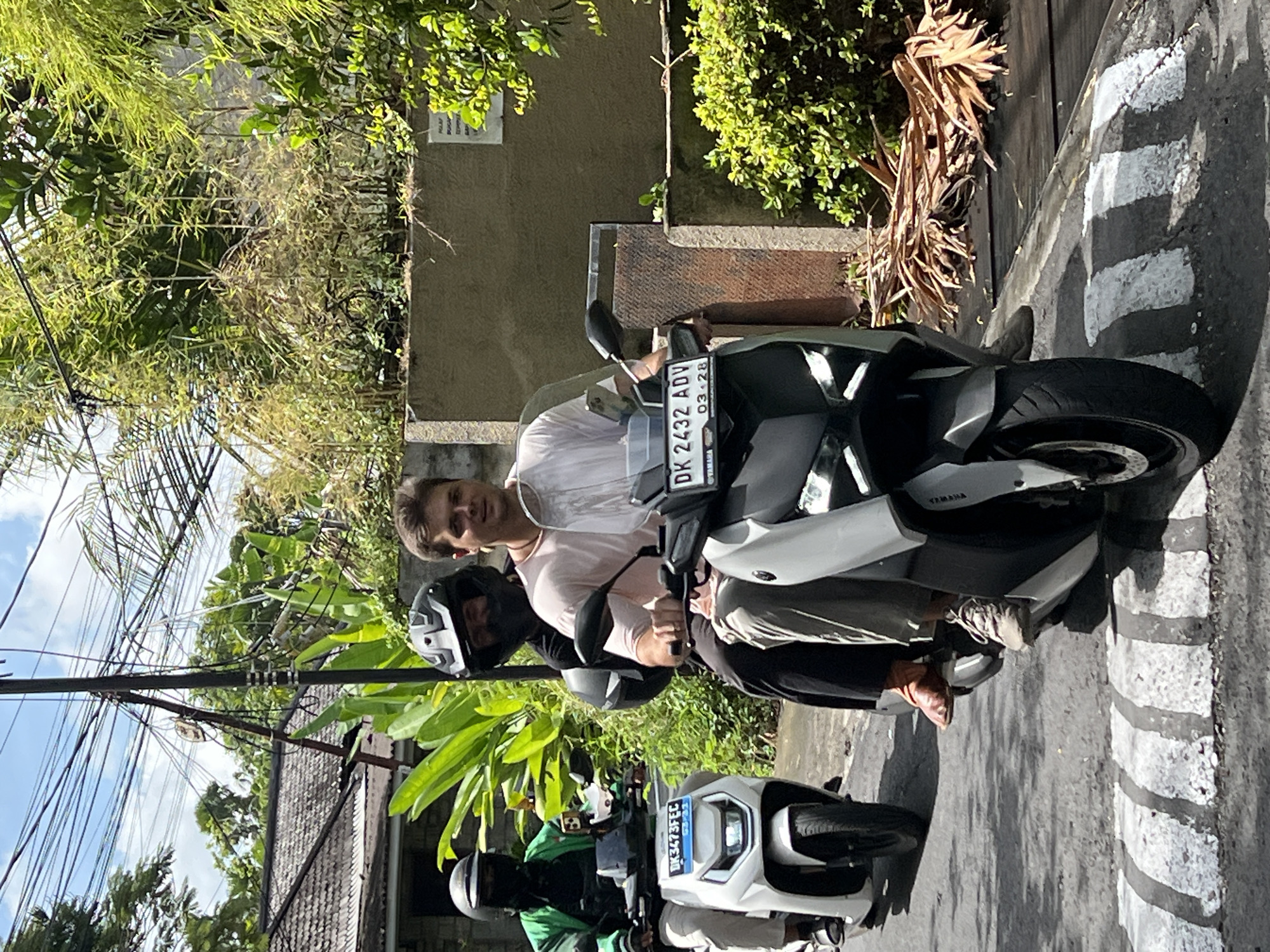 Two individuals, a man and a woman, are riding on a white and gray electric scooter in North Kuta, Indonesia. The man, wearing a light pink shirt, is driving the scooter and is seated in front. The woman, wearing a black shirt and a black helmet, is the passenger behind him, also wearing a helmet. A second scooter, white in color, is to their left, with a person in a green jacket and a black mask riding it. Both scooters have license plates, with the lead scooter displaying "DK 2432 ADV 03.28" and the second scooter showing "DK 3473 FEC 03.23". The scene appears to be a street with tropical vegetation and a gray wall in the background. The road has a speed bump with white stripes.
