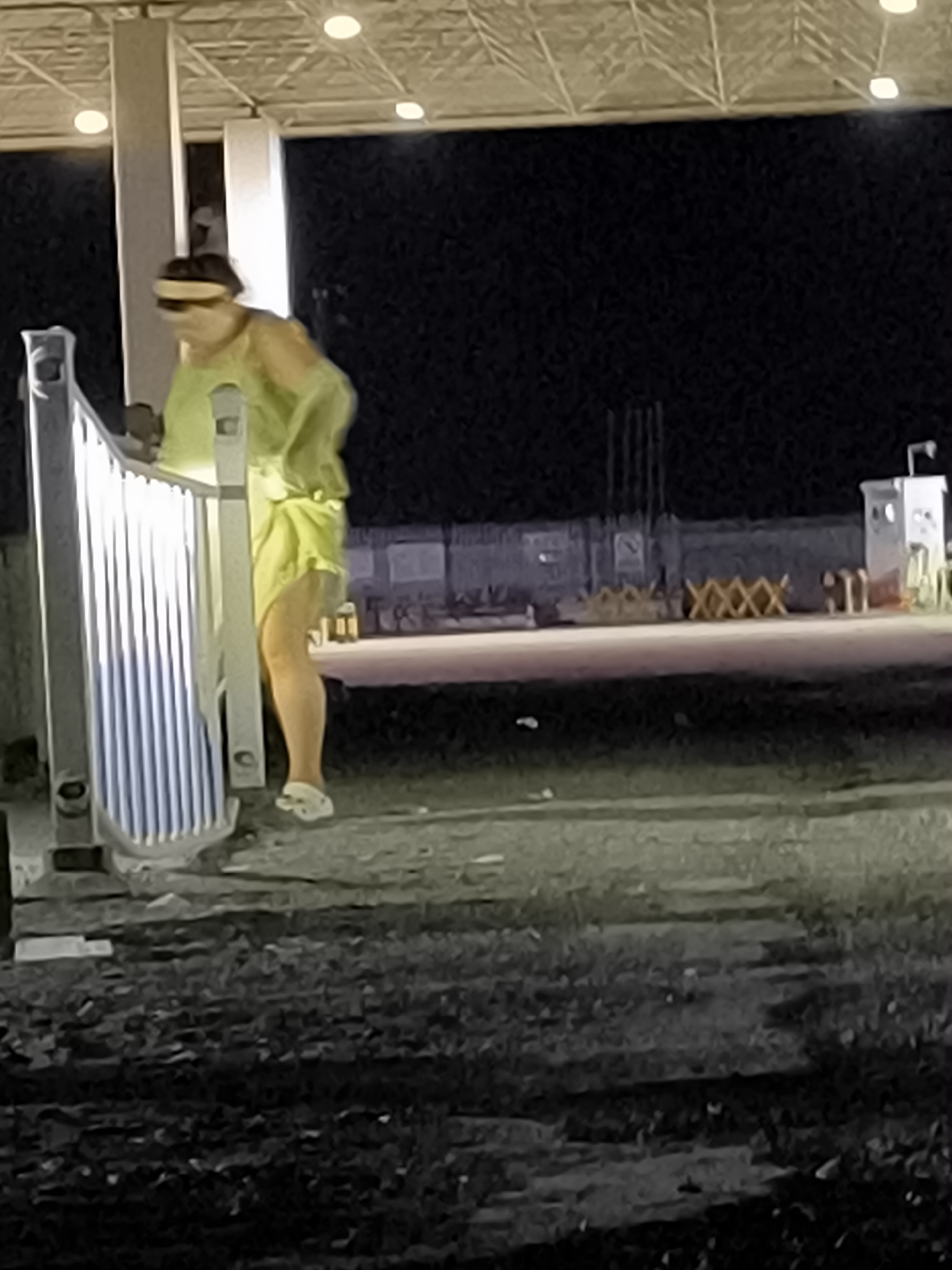 A person wearing a yellow top and white Crocs stands near a white metal barrier in Gurtu Town, China. The person has a yellow band on their head. The scene appears to be outdoors at night, possibly at a construction site or industrial area, with a metal roof structure and lights visible in the background. There are also some barriers and structures in the distance. The ground is uneven and covered with dark material.