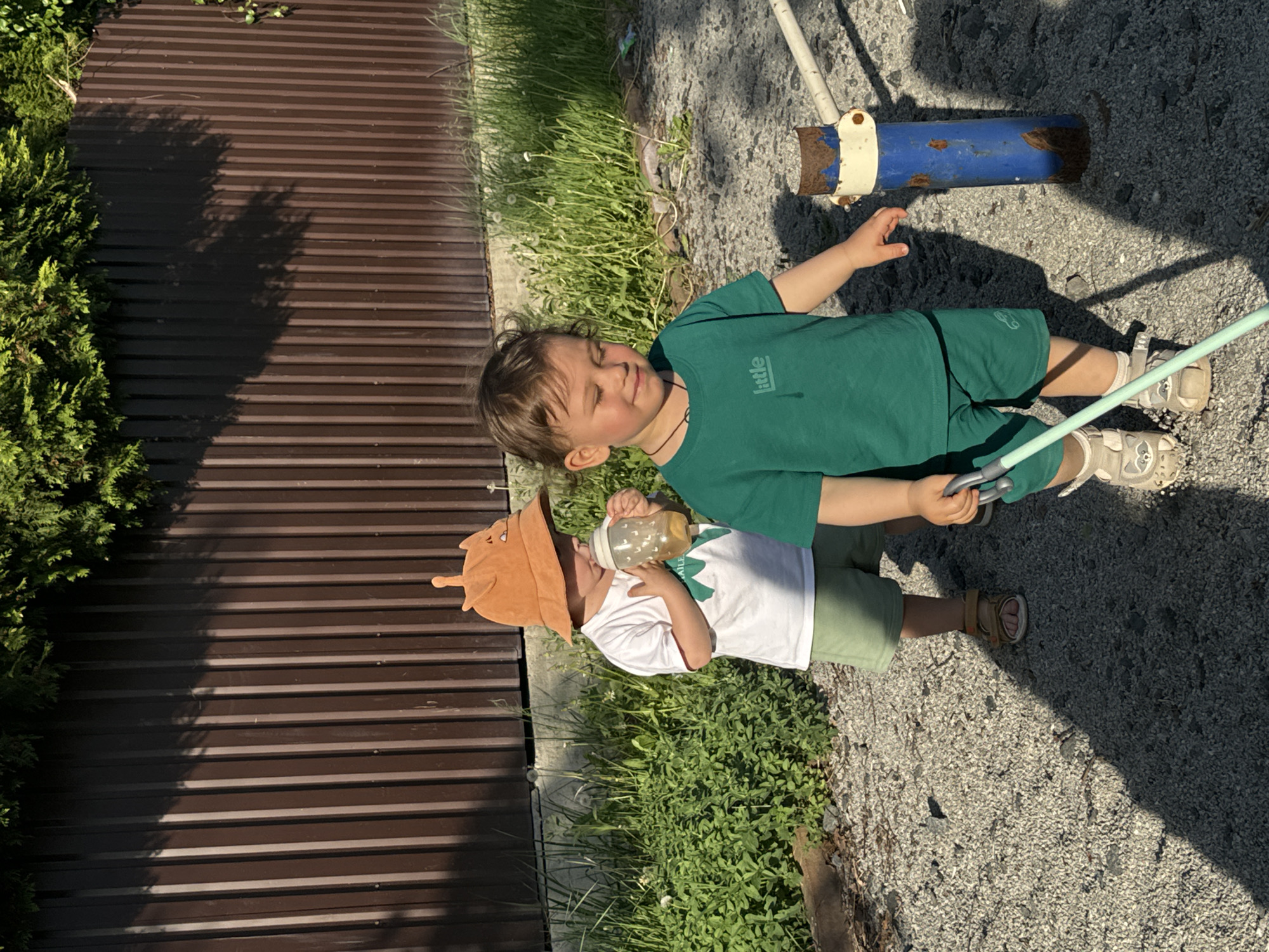 In Селекционер-2 Снт, Russia, two children are outdoors on a sunny day. One child, wearing a white shirt, green shorts, and a brown hat with antennae, holds a bottle. The other child is dressed in a green shirt and green shorts, and holds a fishing rod. They are standing on gravel near a metal fence and vegetation. A blue and white metal object is also visible in the background.