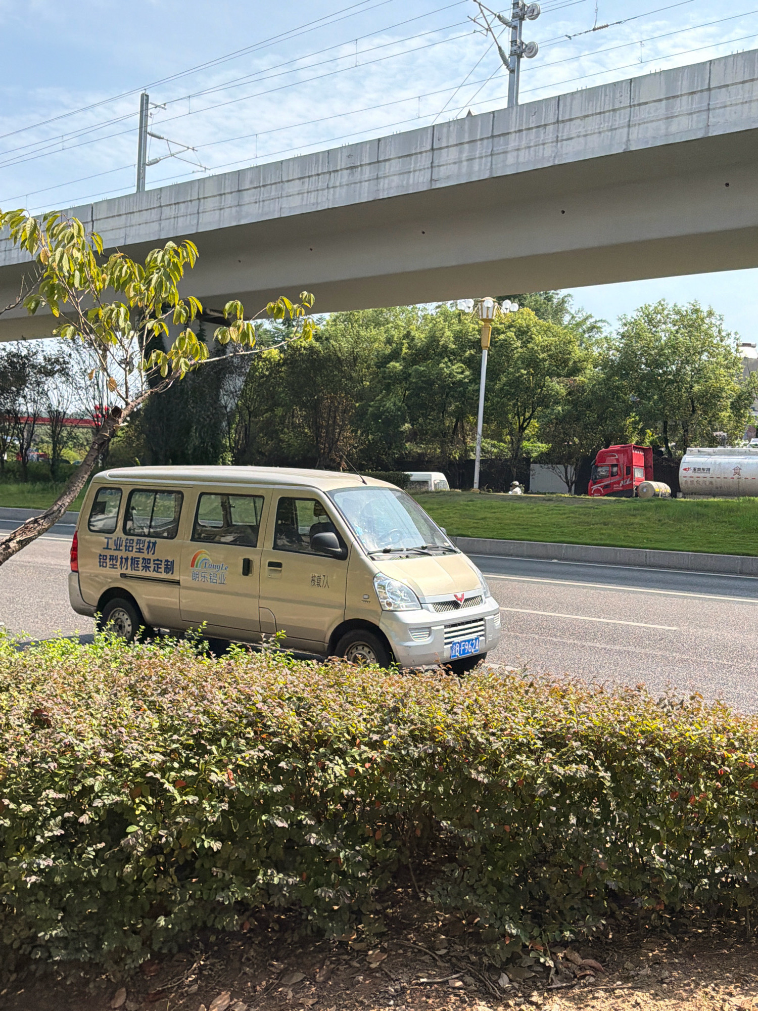 A gold van is parked on the right side of a two-lane road, partially obscured by a dense, green bush in the foreground. The van has Chinese text and a rainbow logo on its side. A tree with green leaves is visible to the left of the van. In the background, a raised concrete structure, possibly an overpass for a train, runs horizontally across the image. Beyond the overpass, more trees and vegetation are visible, along with a red truck and a white tanker truck. The sky is blue with some clouds. The scene appears to be in an urban or semi-urban environment.