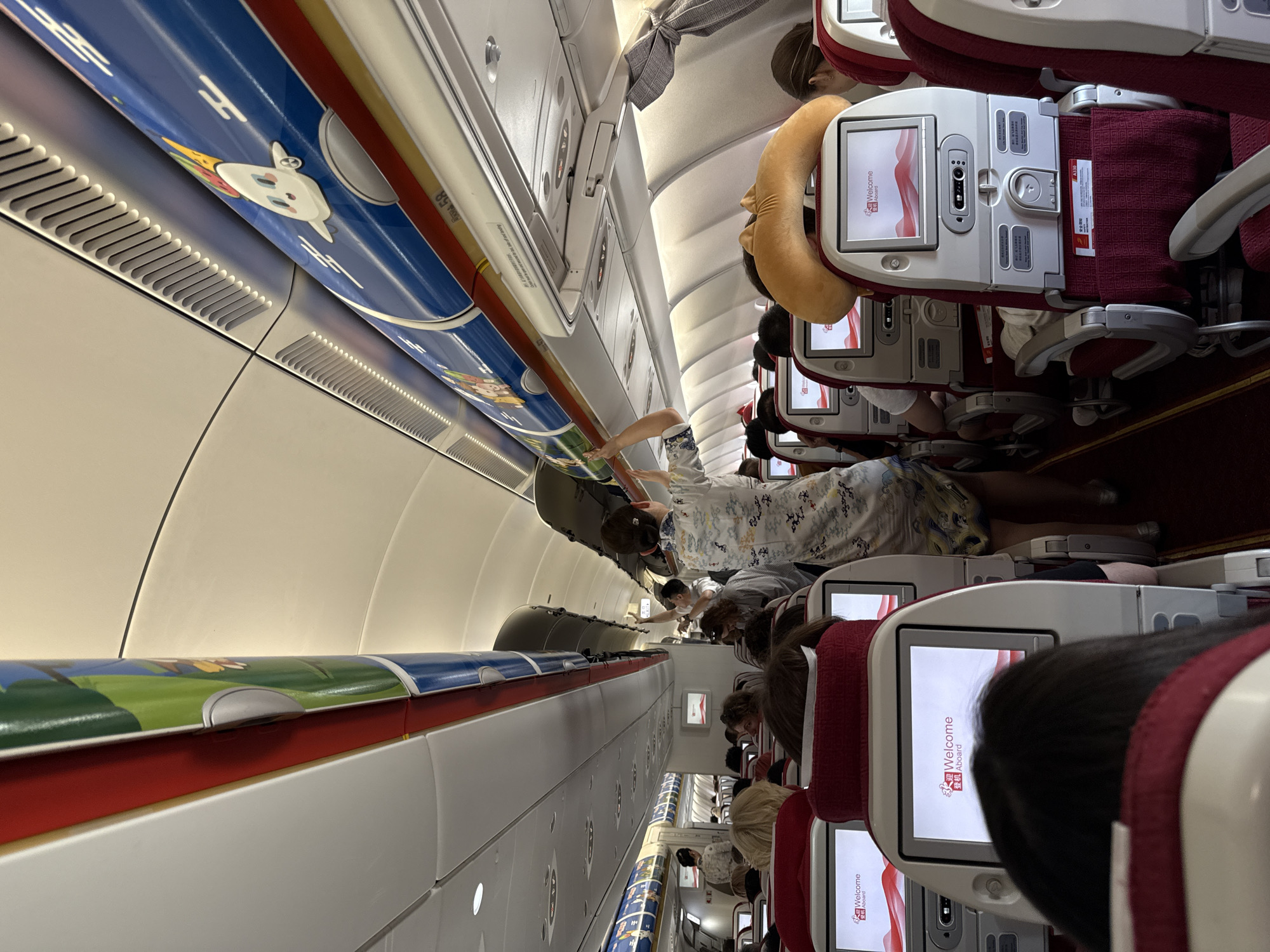 The image depicts the interior of an airplane cabin, viewed from the perspective of a passenger seated in the aisle. The overhead compartments are decorated with cartoon characters and designs. Passengers are seated in rows of red-upholstered seats, many with individual entertainment screens displaying a "Welcome Aboard" message. In the aisle, a flight attendant in a patterned dress is reaching into an overhead compartment, while another flight attendant in a white uniform stands further down the aisle. Some passengers are visible in their seats, and one person in the foreground has their back to the camera. The overall environment suggests a commercial flight. The presence of "Xiaoshan District" in the prompt suggests a potential location for the Xiaoshan International Airport (HGH), which serves the Hangzhou metropolitan area, including Xiaoshan District.