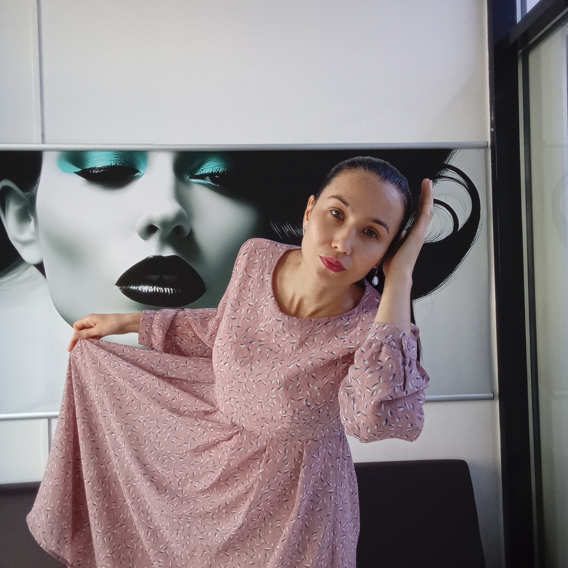 A woman stands in front of a large poster in what appears to be an indoor setting. She is holding the side of her long pink dress with a leaf pattern. Her hair is pulled back, and she is wearing earrings.
The poster behind her features a black and white image of a woman's face with prominent glossy black lips and turquoise eyeshadow. The environment is a room with what looks like a black chair or bench partially visible in the lower part of the frame. A window frame is visible to the right of the poster.
While specific details suggest it might be a beauty salon or related establishment, and given the request for the location, it's possible this image was taken in Izhevsk, Russia. However, there are no immediately apparent visual indicators of the city itself or specific landmarks.