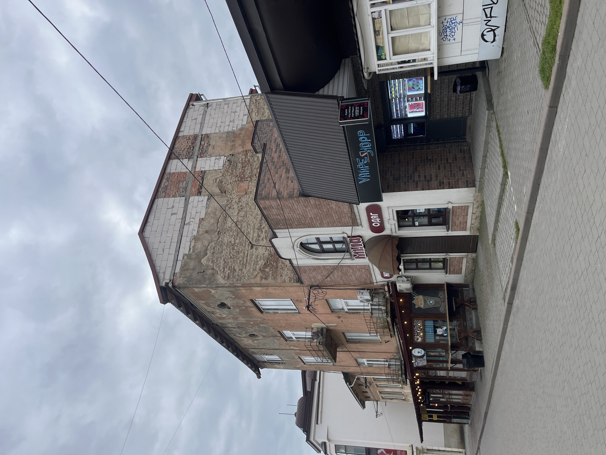 The image shows a street scene in the city of Drohobych, Ukraine. The foreground is a paved street and sidewalk. On the right side of the street is a white kiosk with graffiti. Next to it is a vape shop with a dark awning and a sign that reads "VAAPE SHOPP". A window displays various vape products. To the left of the vape shop is a building with a pinkish facade, showing signs of disrepair, with balconies and air conditioning units. Above this building, an unfinished upper level made of white bricks is visible. To the left of the pink building is a cafe with an awning, outdoor seating, and string lights. Further to the left, another building with white and light-colored walls is partially visible. The sky is overcast.