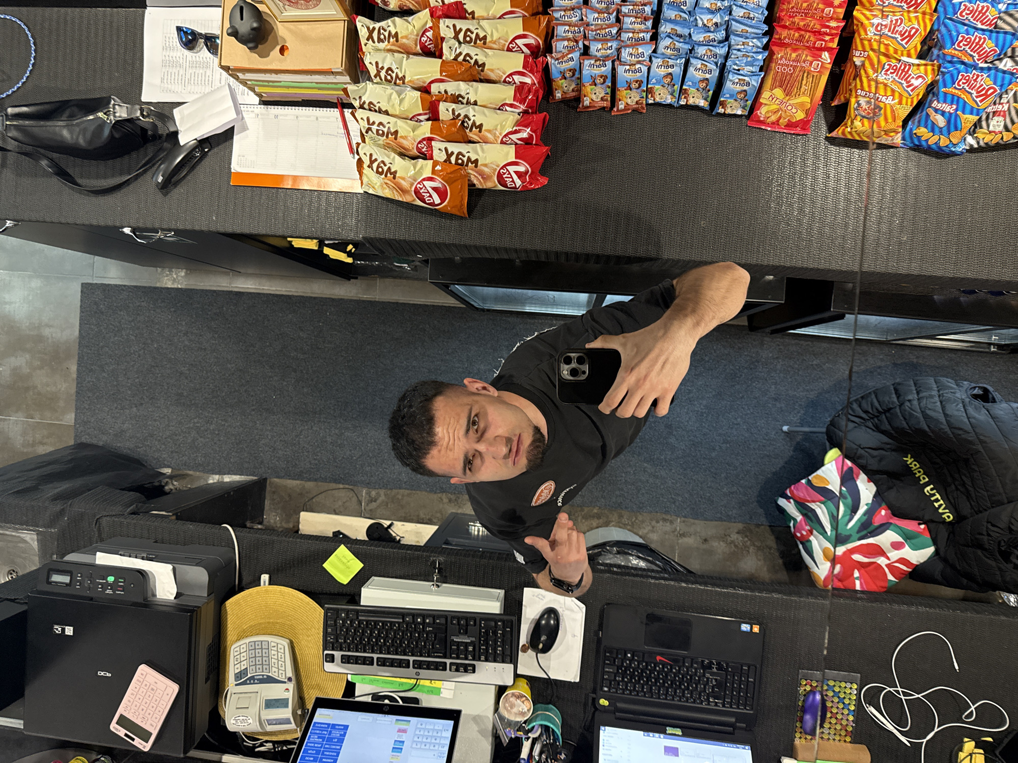 The image shows a man taking a selfie in what appears to be a convenience store or small shop. He is holding a phone and looking directly at the camera. In the background, there is a counter stocked with various snacks, including packages of "X6M" and "BOLU!" brand items. To the left of the man, there is a workspace with a printer, calculator, keyboard, mouse, and laptop. A black jacket and a colorful bag rest behind the counter. The location, Shumen, Bulgaria, is inferred from the brands "X6M" and "BOLU!" which are associated with the region.