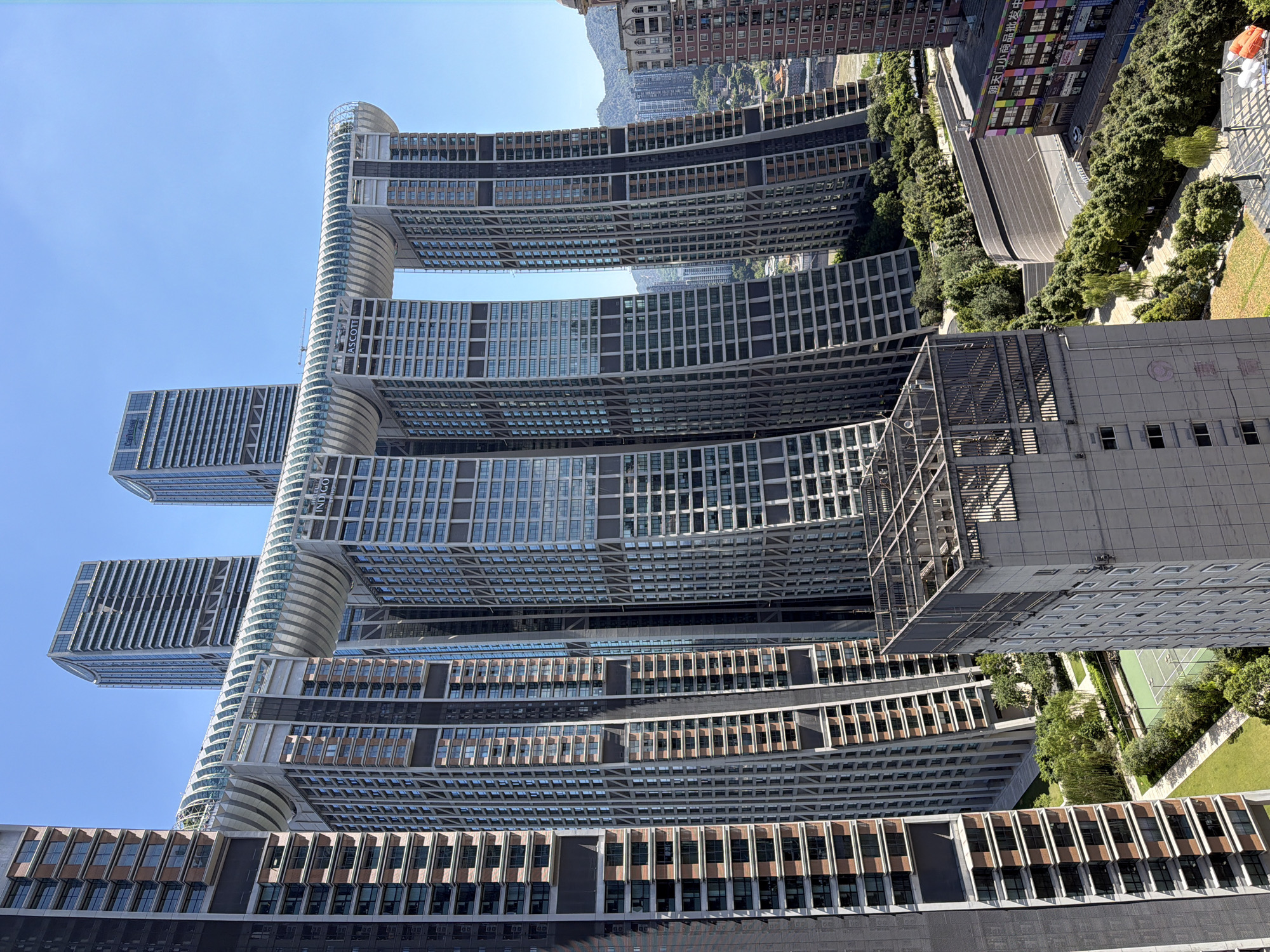 The image captures the unique "Crystal" building complex in Yuzhong District, Chongqing, China, on a clear, sunny day. The complex consists of four skyscrapers connected by a long, curved skybridge that resembles a ship's hull. The buildings have a layered design with staggered setbacks and a facade of glass and concrete. The skybridge is enclosed with glass panels, allowing for panoramic views.
In the foreground, to the left, a portion of a dark-colored skyscraper with a regular pattern of windows and balconies is visible. To the right of the main complex, a smaller, lighter-colored building with a grid-like pattern of windows is partially seen. Below the skybridge and between the buildings, there is a landscaped area with trees, grassy patches, and a tennis court. In the background, more buildings of varying heights, some appearing to be residential, are nestled against rolling hills. A road winds through the lower right section of the image, lined with trees and buildings, including one with colorful signage. The sky is a vibrant, clear blue, indicating good weather conditions. There are no people or visible vehicles in the immediate foreground or on the skybridge.