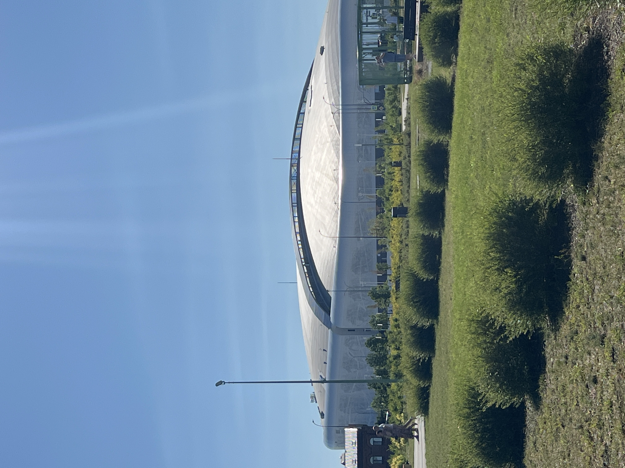 A modern, dome-shaped stadium, likely the Nizhny Novgorod Stadium, is viewed from a grassy area with neatly trimmed bushes in the foreground. The stadium, predominantly silver or white, features a curved roof with reflective panels and a ribbed texture. A tall lamppost stands to the left of the stadium, and a red brick building with multiple windows is partially visible to its left. Two people are walking on a paved path in front of the stadium. To the right of the stadium, a green, open-air enclosure with benches and a person standing inside can be seen. The sky is clear and blue with some light flare, suggesting a bright, sunny day.