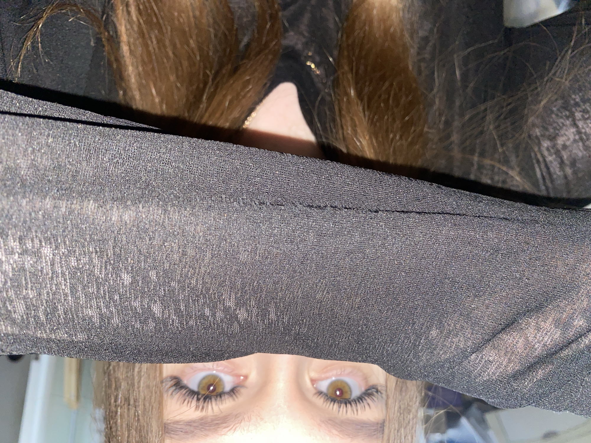 A frontal close-up captures an individual, appearing female, with light brown (hazel) eyes and distinctly long, dark eyelashes. Their left forearm, encased in a black, semi-sheer, finely textured fabric, is held horizontally across the lower portion of their face, obscuring the nose, mouth, and chin. Only the upper part of the face, including the eyes, eyebrows, and forehead, is clearly visible above the arm. Long, light brown hair frames the face and extends below the arm, with some strands visible over the black fabric. A thin, gold-colored chain necklace is faintly visible at the subject's neck, beneath what appears to be a dark, possibly black, inner garment. The background is significantly out of focus, rendering specific details indistinguishable, though it appears to be an interior space with diffuse lighting. The image provides no discernible information regarding its location in the city of Chaikovsky, Russia, or any other specific geographical area.
