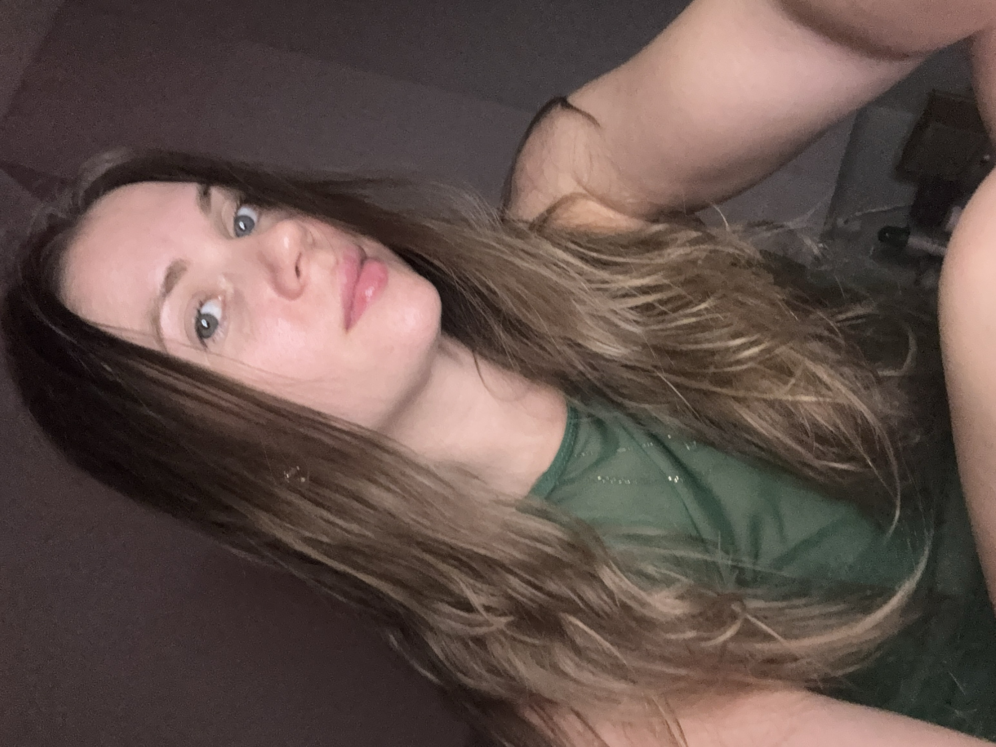 A woman is lying down, likely in an indoor environment with dim lighting. She has long brown hair and blue eyes. She is wearing a green top. The background is blurred, obscuring details of the location. There is no information about this image being taken in the city of Batumi, Georgia.