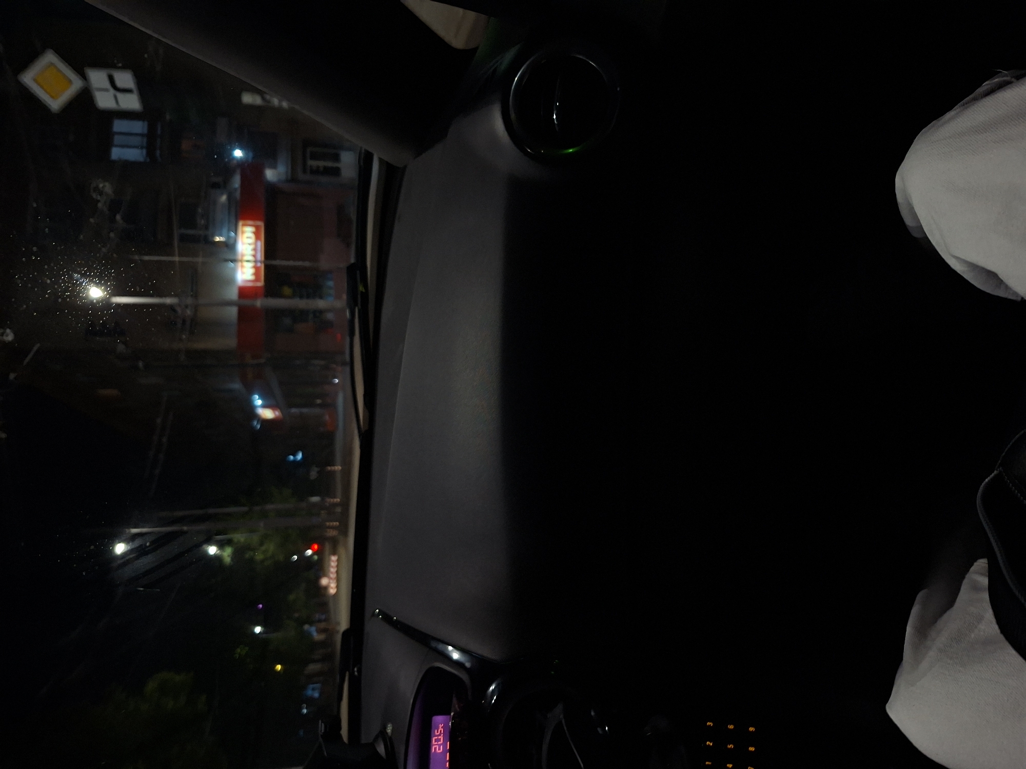 The image, taken from the interior of a car at night, shows a view through the windshield of a city street. The street is wet, reflecting lights from streetlamps and building signs. Two traffic signs are visible on the left, including a yellow diamond-shaped sign and a white sign with a black arrow. Across the street, a tall red illuminated sign with "NORDI" is prominent on a building.
Inside the car, part of the dashboard is visible, including a display showing "20.5°C". There's a circular vent on the dashboard. A person's sleeve is seen on the right edge of the photo.
Based on the "NORDI" sign, it is highly likely that the location is Pleven, Bulgaria.