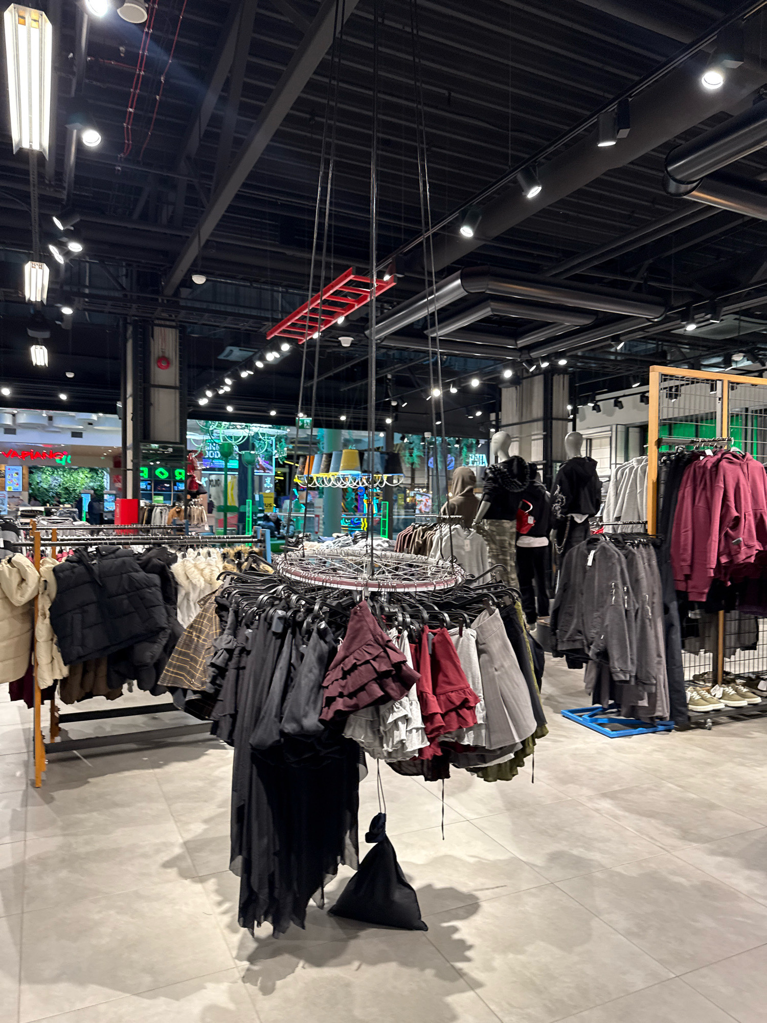 The image depicts the interior of a clothing store in Tallinn, Estonia. The store features a modern, industrial aesthetic with exposed ceilings, metal beams, and track lighting. Clothing racks filled with various garments, including jackets, dresses, and sweaters, are prominently displayed. Some mannequins are visible in the background, showcasing outfits. The floor is tiled in a light gray color. The overall impression is one of a well-lit, organized retail space, likely a fashion boutique or department store.