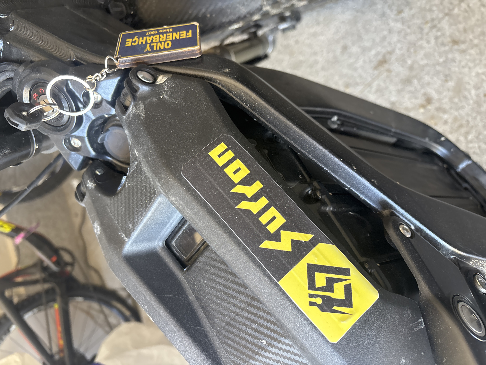 The image shows a portion of a black Sur Ron electric motorcycle. The motorcycle has a "Sur Ron" sticker on the frame. Keys are in the ignition, along with a keychain that reads "ONLY FENERBAHCE Since 1907". A bicycle is partially visible in the background. The location is not identifiable as Asenovgrad, Bulgaria.