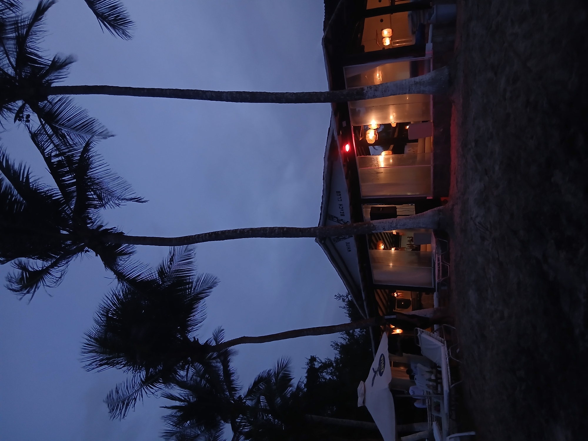 At twilight in Playa Parguito, Venezuela, a two-story beachfront structure with transparent walls emits light from within. It has a sign saying "White Beach Club." Palm trees are visible in the left foreground, silhouetted against the dusk sky. Beach chairs and a white umbrella stand near the building on the sand. The ground in the right foreground is dark.