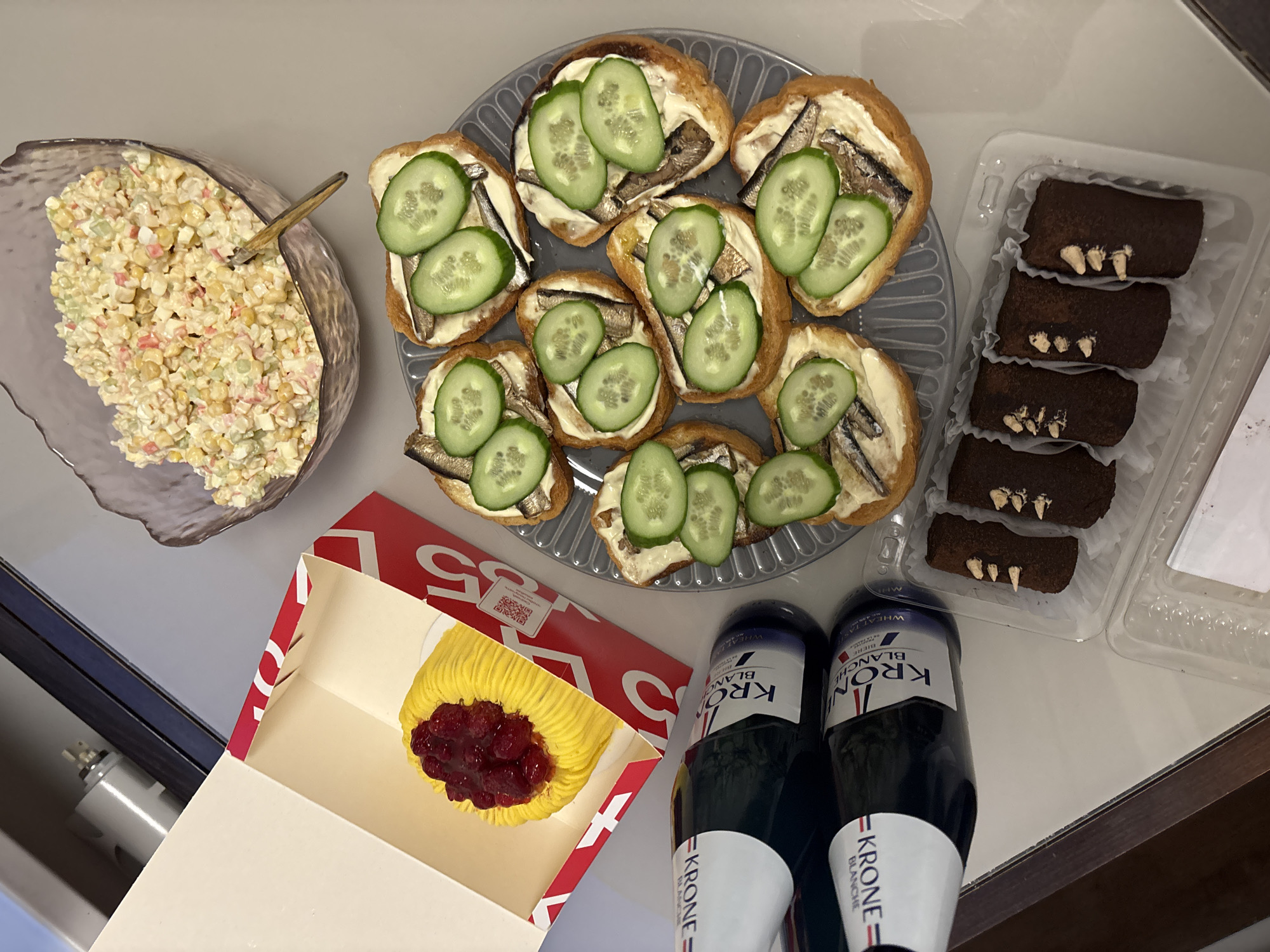 A high-angle shot depicts a spread of food and drink items on a light gray surface. Clockwise from the top left, there's a glass bowl filled with a corn-based salad containing visible crab sticks and a spoon resting inside. Next, there's a clear gray plate with approximately eight sandwiches. The sandwiches appear to have a white spread, small fish, and cucumber slices on top. To the right of the sandwiches are six rectangular chocolate cakes in a clear plastic container. Moving down, there's an open box revealing a single yellow cake topped with red berries. Positioned near the cake box are two dark-colored bottles, labeled "KRONE BLANCHE". A corner of what may be a window frame can be seen on the lower left side of the image. The overall composition suggests a casual gathering with a variety of food and drink options.
Based on food items, beverages and other clues in the image, location of this image may be inferred to be Saint Petersburg, Russia.