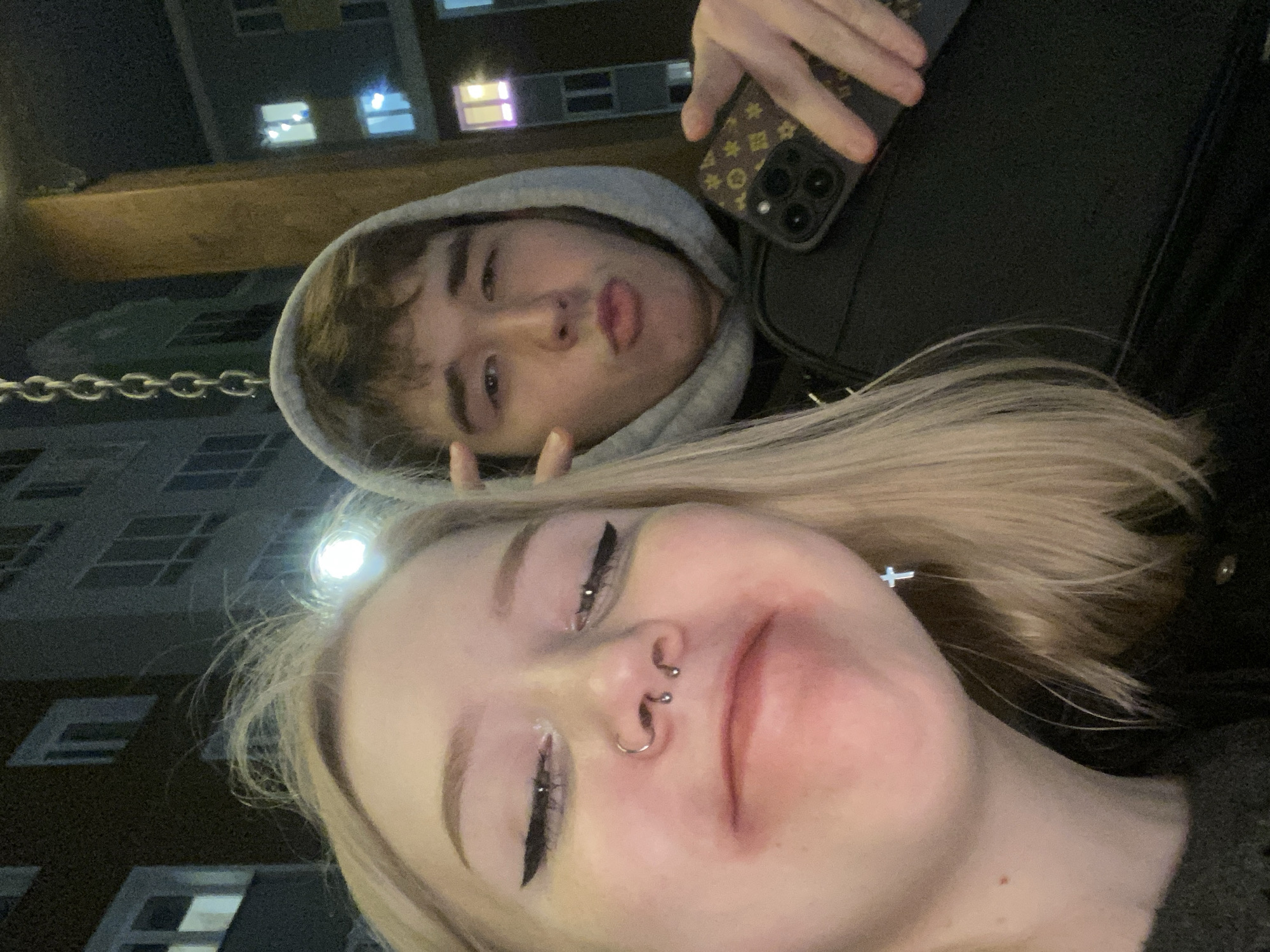 Two people are posing for a selfie, with the person with blonde hair closest to the camera and the other person wearing a grey hooded sweatshirt behind them. The person in the hooded sweatshirt is making a duck face and holding up two fingers. The blonde-haired person has multiple nose piercings and is wearing a cross earring. A black phone case with a brown and gold design is visible in the upper right corner. The background shows a multi-story building with lit windows, suggesting it is nighttime. The photo was taken outdoors, possibly on a swing due to the visible chain. The location is not Pervomaysky, Russia.