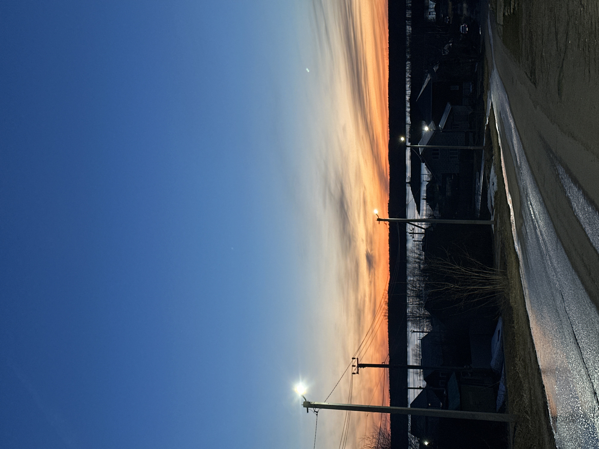 The image captures a twilight scene in Chaikovsky, Russia. The sky transitions from a deep blue on the left to shades of orange and pink near the horizon. A sliver of the moon is visible in the upper center of the sky. Below the horizon, the silhouettes of trees, houses, and utility poles are visible. Streetlights illuminate a wet road on the right side of the frame, reflecting the ambient light. No people are visible.