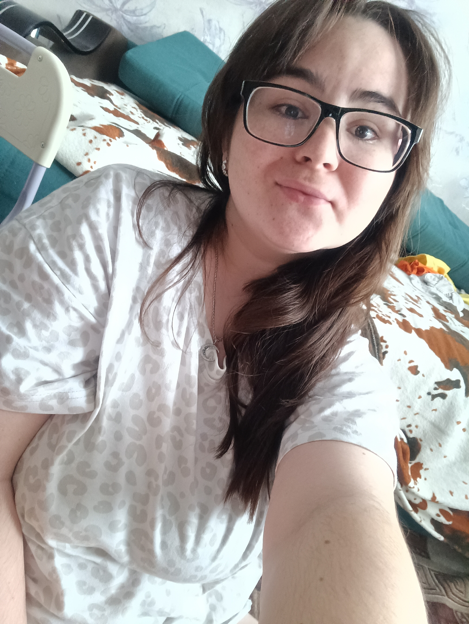 A selfie taken in an indoor environment featuring a woman with brown hair and glasses. She is wearing a patterned shirt. Behind her, there's a patterned blanket, a blue cushion, and a partially visible baby high chair. The location is Noviy Urengoy, Russia.