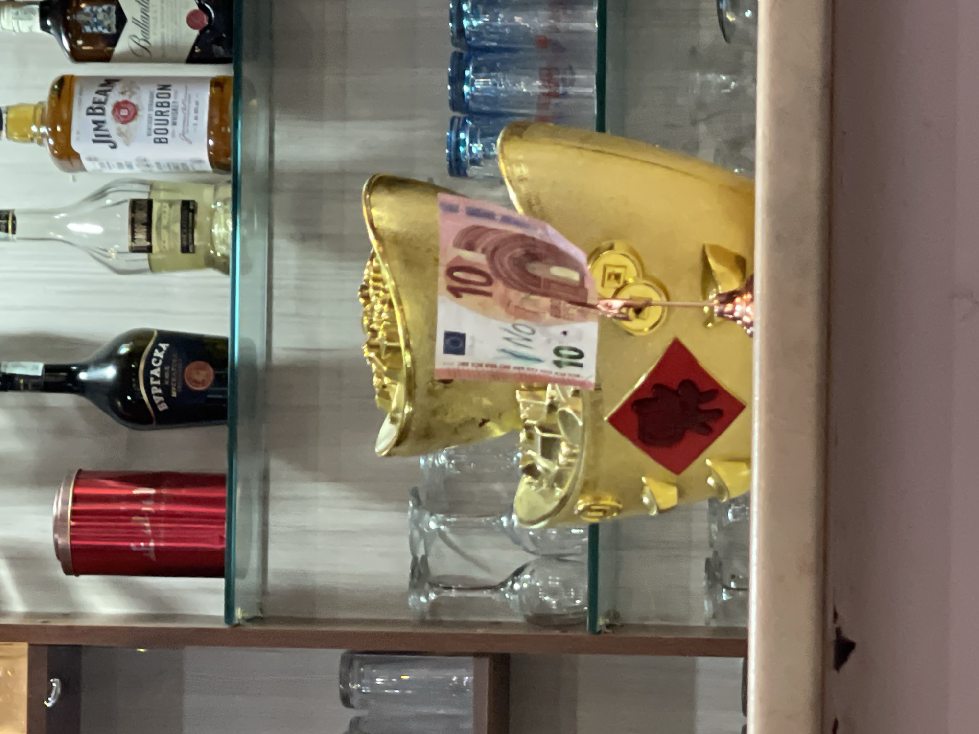 The image depicts a shelf, likely inside a cabinet or display case, containing an assortment of items. On the upper shelves, there are several bottles of alcoholic beverages including bourbon and vodka. Below the alcoholic drinks, there is a red container, potentially a beverage can or a cylindrical tin. In front of these items on the shelf, there is a gold-colored decorative piece resembling an ingot, adorned with traditional Chinese elements such as coins and a red square with a symbolic motif. A 10 Euro banknote is clipped to the ingot. The setting appears to be indoors, possibly a shop or restaurant in Sofia, Bulgaria, given the presence of the Euro currency. Additionally, there are several glasses, indicating possible bar location. No people are visible in the image.