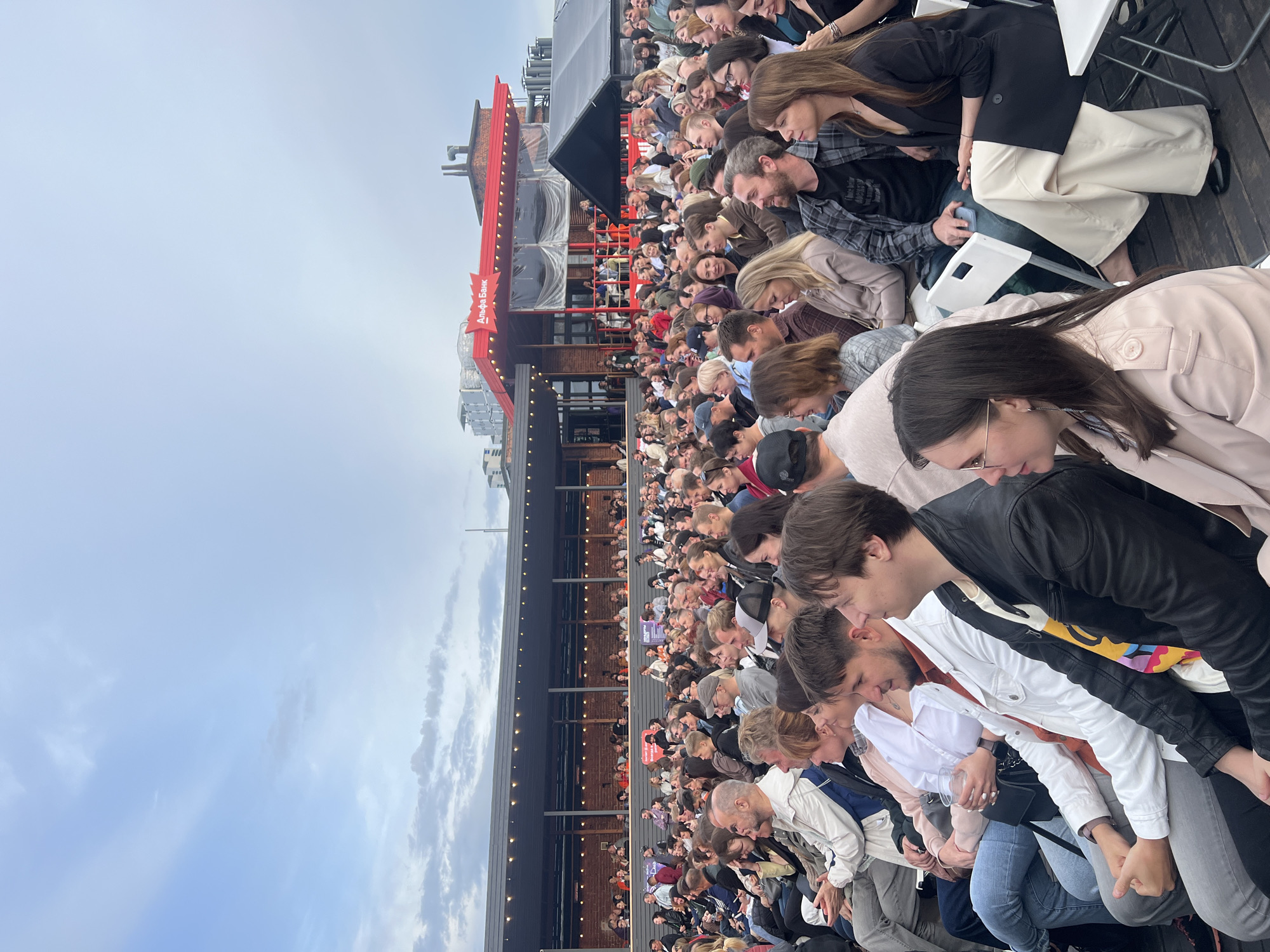 The image shows a large crowd of people gathered outdoors, seated on what appears to be a tiered seating area, possibly a stadium or outdoor event space. The perspective is from behind the crowd, looking down and towards the left. The people are mostly facing away from the camera, focused on something to their left. They are dressed in casual attire, suggesting a relaxed, informal event.
The scene is set against a backdrop of a cloudy sky, indicating it is likely daytime, though the weather appears overcast. To the left of the frame, there is a red building with a sign that has Cyrillic text. Part of the sign is visible and reads "Альфа Банк," which translates to "Alfa-Bank," a Russian bank. This text, along with the setting, suggests the location is Saint Petersburg, Russia. The overall mood appears to be one of anticipation or engagement, as the crowd is intently watching something out of frame. The lighting is diffused due to the overcast sky.