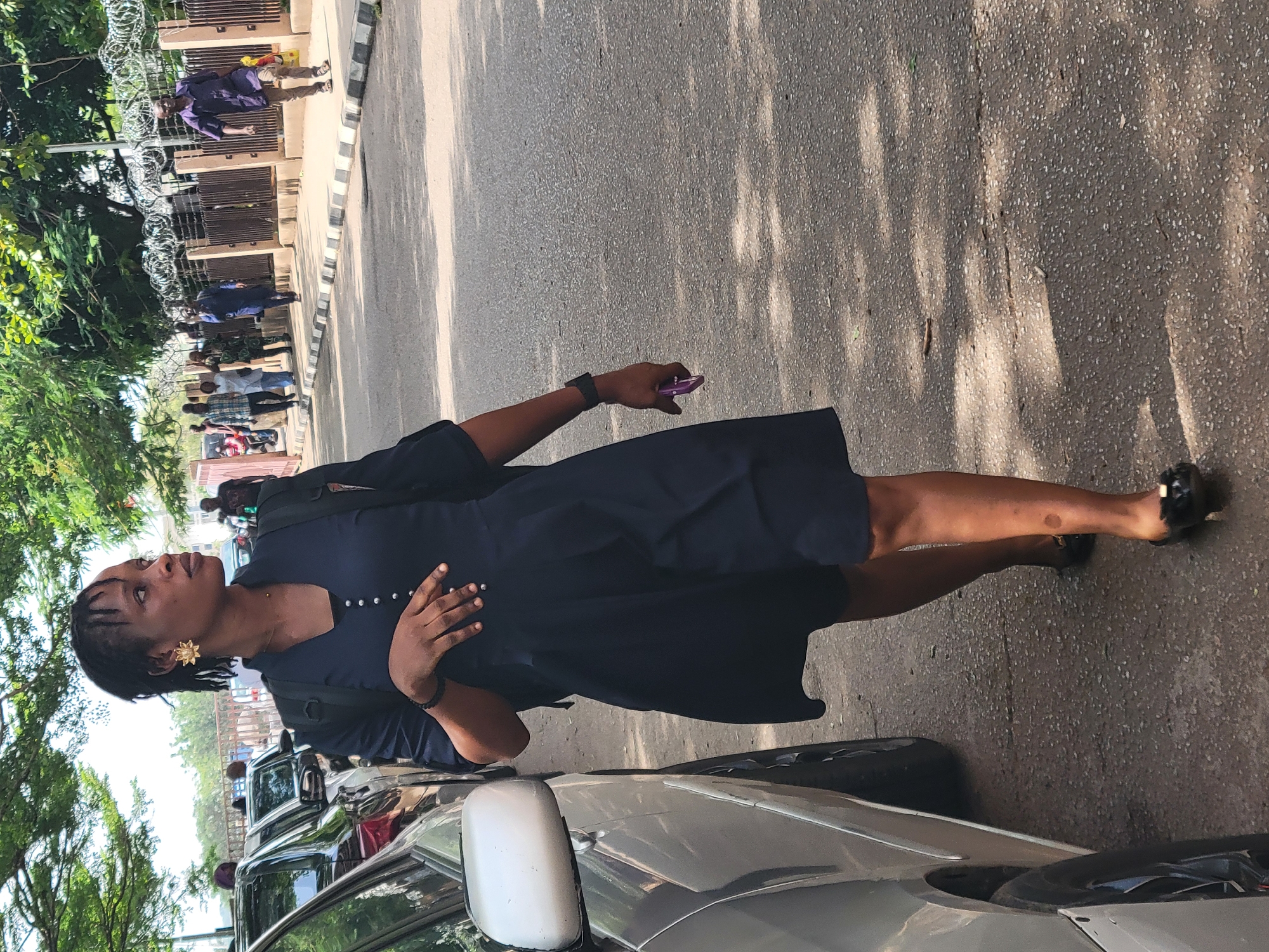 A woman is walking along a street in Abuja, Nigeria. She is wearing a navy blue dress, black flats, a backpack, and gold-colored earrings. She is holding a purple cell phone in her right hand and has a dark bracelet on her left wrist. The street is lined with parked cars on the left and a sidewalk with a metal fence on the right. Other people are visible in the background near the fence. There are trees and foliage visible in the background and the foreground of the image. Shadows are cast on the street.