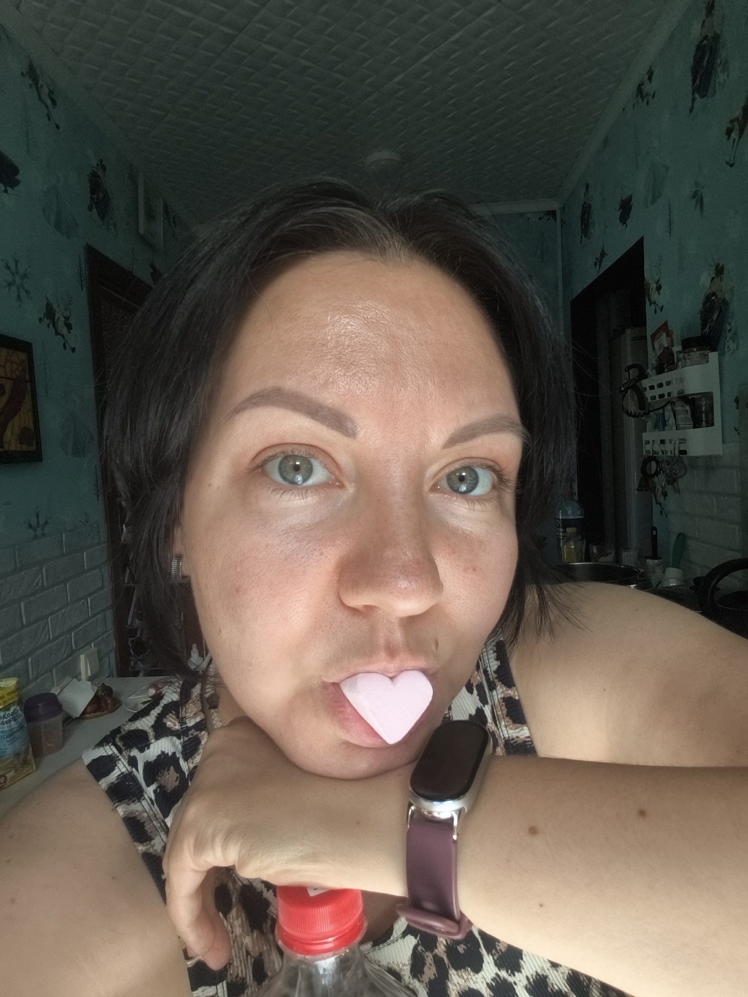 A close-up, eye-level, indoor selfie shows a woman with short, dark hair and light skin holding a pink, heart-shaped object in her mouth. Her eyes are open, and she is looking directly at the camera. She is wearing a black and white leopard-print top and a dark-colored smartwatch on her left wrist. Her left arm is resting on a bottle with a red cap. The background consists of a teal-colored wall with a patterned wallpaper and a textured ceiling. Shelves with various items are visible on the wall to the right. The overall environment appears to be a domestic setting, likely a kitchen or living area. There are no discernible landmarks to pinpoint the location to Orenburg, Russia.