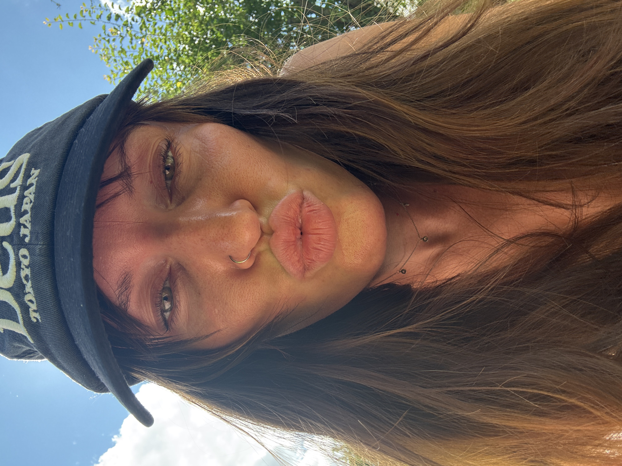 A woman with long brown hair and a nose ring puckers her lips for a selfie. She is wearing a black visor with white lettering that reads "DEUS TOKYO JAPAN." The background shows a bright blue sky with white clouds and green foliage, suggesting an outdoor setting. The lighting indicates it is daytime. The image is a close-up, focusing on the woman's face and upper body. There is no indication of the location being Красная Пресня Снт, Russia.